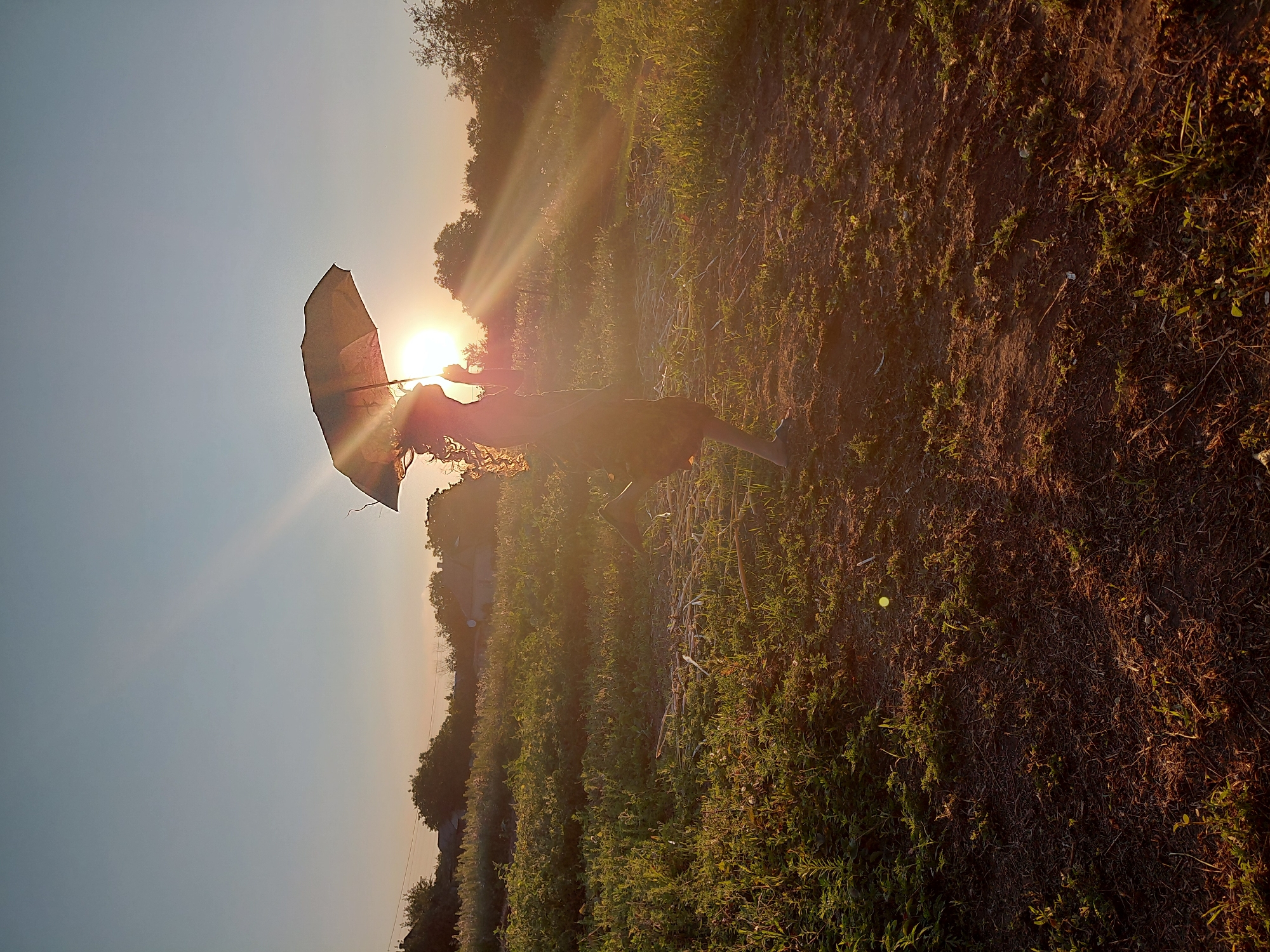 The image, set in a rural outdoor location, captures a silhouette of a young person with curly hair holding a dark umbrella. The person is mid-leap, with one leg extended forward and the other bent behind. They are wearing a dark, possibly flowy dress that extends below the knee. The sun is low on the horizon, casting long rays of light and creating a halo effect around the person's head and umbrella. The foreground is a patch of dry, brown earth with sparse green vegetation, while the middle ground features more lush green plants and some dry stalks. In the background, trees and the roofs of houses are visible against a pale blue and orange sky. The scene appears to be a field or a backyard during sunset. The location is identified as Unknown, Unknown.