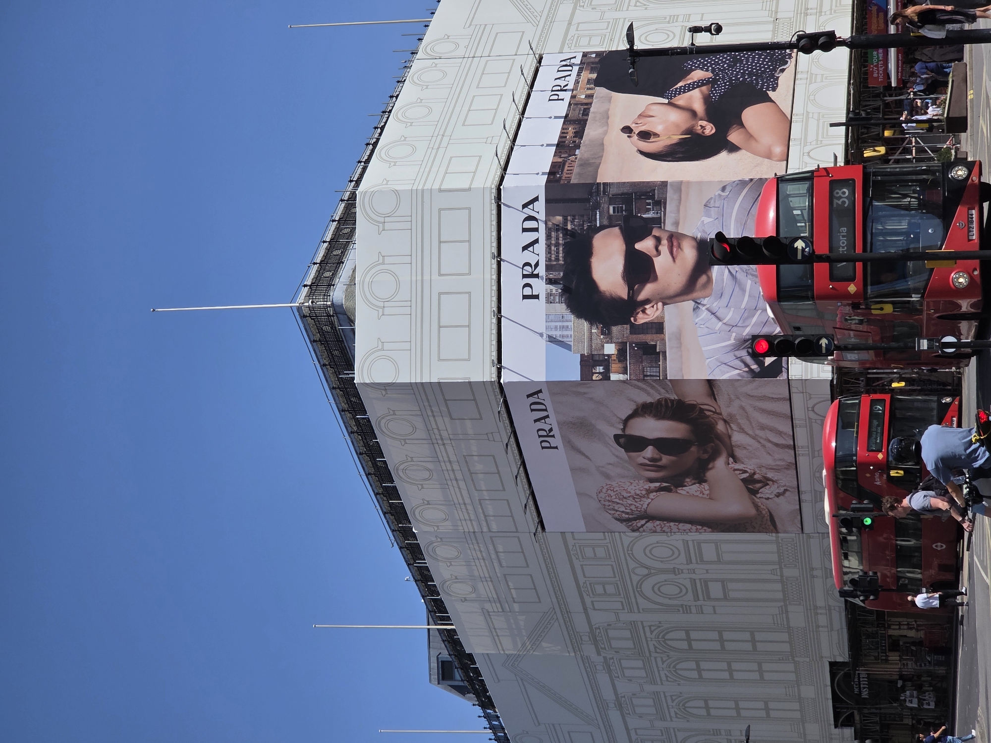 The image shows a building facade covered with Prada advertisements in London, United Kingdom. There are three large, vertically stacked advertisements featuring models wearing sunglasses. A red double-decker bus passes by on the street, specifically Route 38 (Victoria). There is also a Route 19 red double-decker bus and a red traffic light. People are present on the sidewalk and on the bus. The sky is bright blue. A Piccadi Institute sign is visible at the bottom of the building.