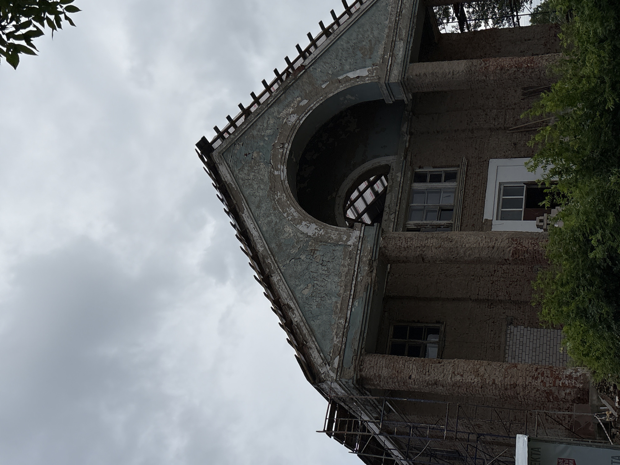 The image shows the exterior of a dilapidated building, likely undergoing renovation or demolition, under a cloudy sky. The building features a large, arched facade with peeling blue paint and exposed brickwork. Scaffolding is visible on the left side of the structure. Several windows are present, some boarded up and others broken. Dense green foliage obscures the lower portion of the building and the ground. The scene appears to be located in the city of Kstinin, Russia.