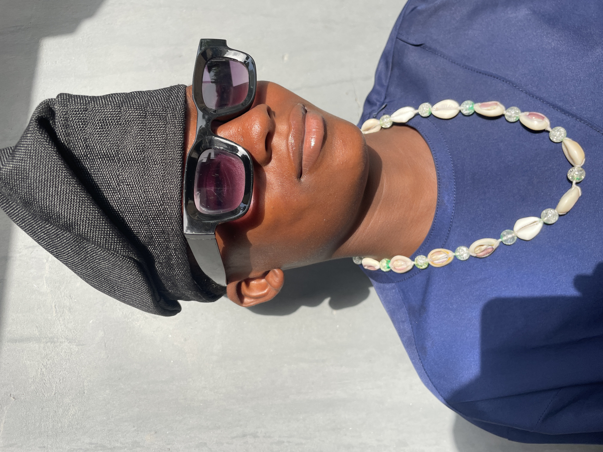 The image displays an overhead close-up of a person with dark skin, seen from the neck up, in a relaxed, reclining pose outdoors. Large, black, rectangular sunglasses with purple-tinted lenses rest on their forehead, obscuring their eyes. A black, textured head covering or hood, exhibiting a subtle woven pattern, is worn underneath the sunglasses. The person's lips are slightly parted.
Around their neck, a necklace made of white cowrie shells alternating with clear, sparkly beads, some of which have a greenish tint, is visible. They are wearing a plain blue t-shirt or top.
The setting features a light-colored, possibly concrete or tiled surface that is brightly illuminated by direct sunlight, casting strong, distinct shadows. This indicates a sunny day, likely during mid-day or early afternoon. The scene evokes a sense of calm and leisure in an outdoor environment, fitting the location context of Badagry, Nigeria. There is no visible text in the image.
