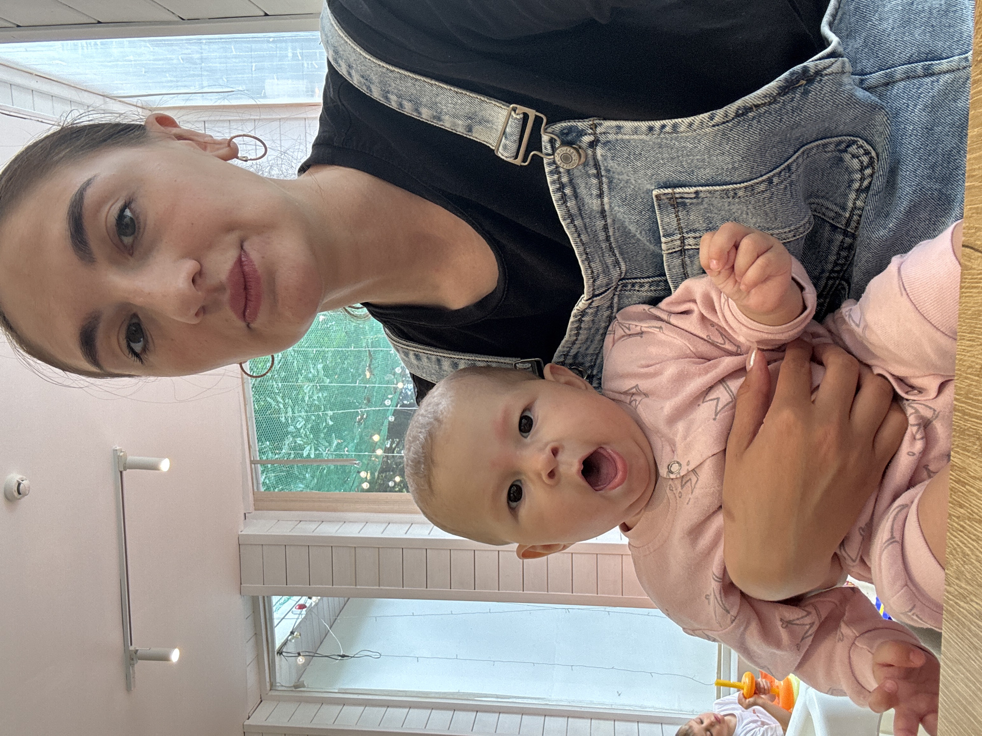 A woman is holding a baby. The woman is wearing a black shirt and blue denim overalls. The baby is wearing a pink outfit. The baby's mouth is open. They are indoors. The environment appears to be a home. The location is not discernable as Sochi, Russia based solely on visual elements of the image.