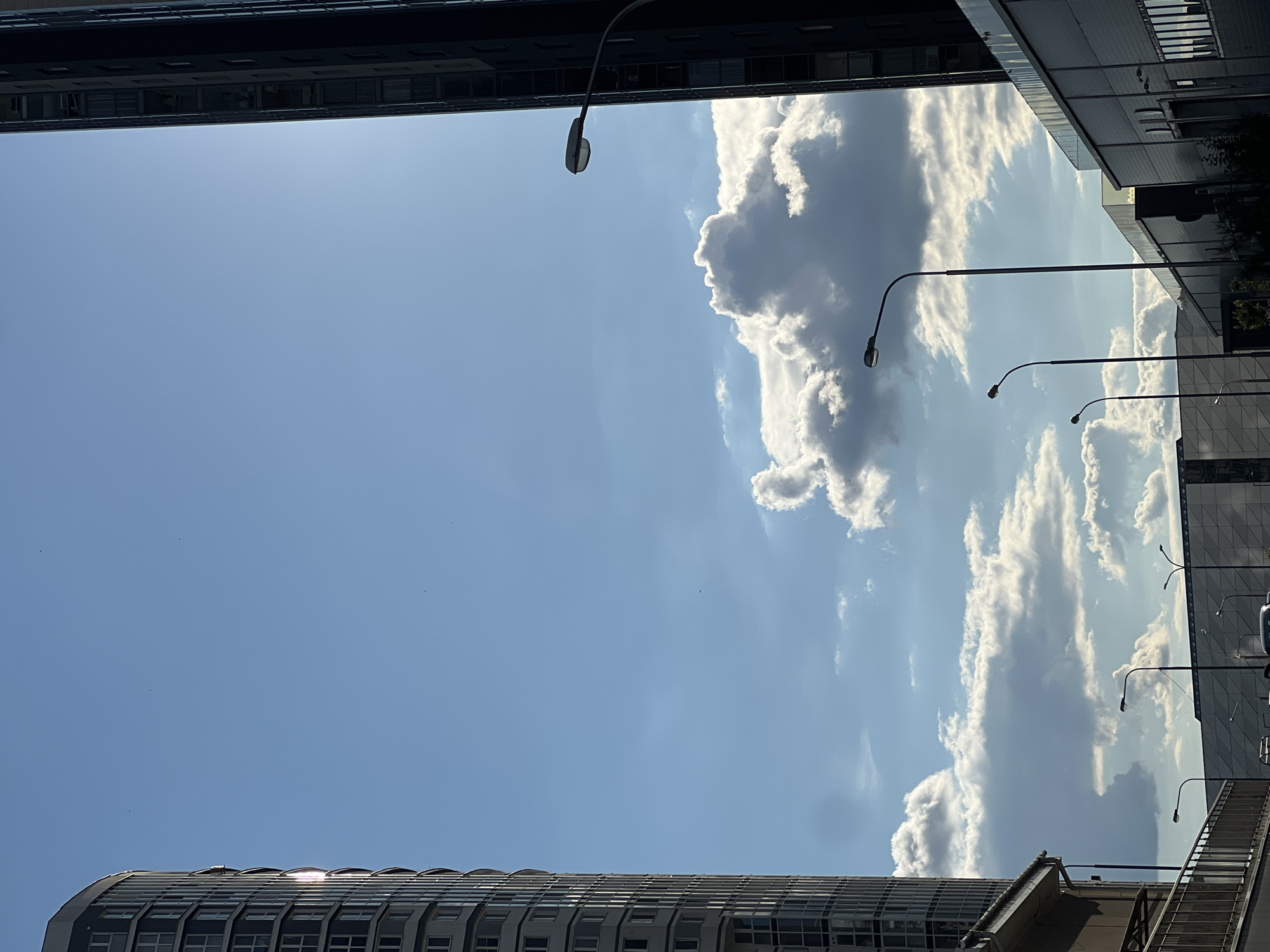 This image, taken on a bright day in Minsk, Belarus, captures a section of the city with a focus on the sky and modern architecture. On the left side, a tall, curved residential building with many windows dominates the frame, its facade reflecting the sunlight. To the right, another, darker, taller building is partially visible, its sleek, modern design contrasting with the lighter building.
The sky is a prominent feature, filled with scattered, fluffy white clouds against a clear blue backdrop. Sunlight streams down, creating a bright atmosphere and highlighting the textures of the clouds and buildings. Several streetlights with curved arms are positioned along the foreground and midground, their dark silhouettes standing out against the sky and the modern, geometric facades of the buildings. These buildings appear to be commercial or office spaces, characterized by their flat roofs and windowed exteriors. The overall scene suggests an urban environment with contemporary architecture and a pleasant, sunny day. There are no people visible in the image.