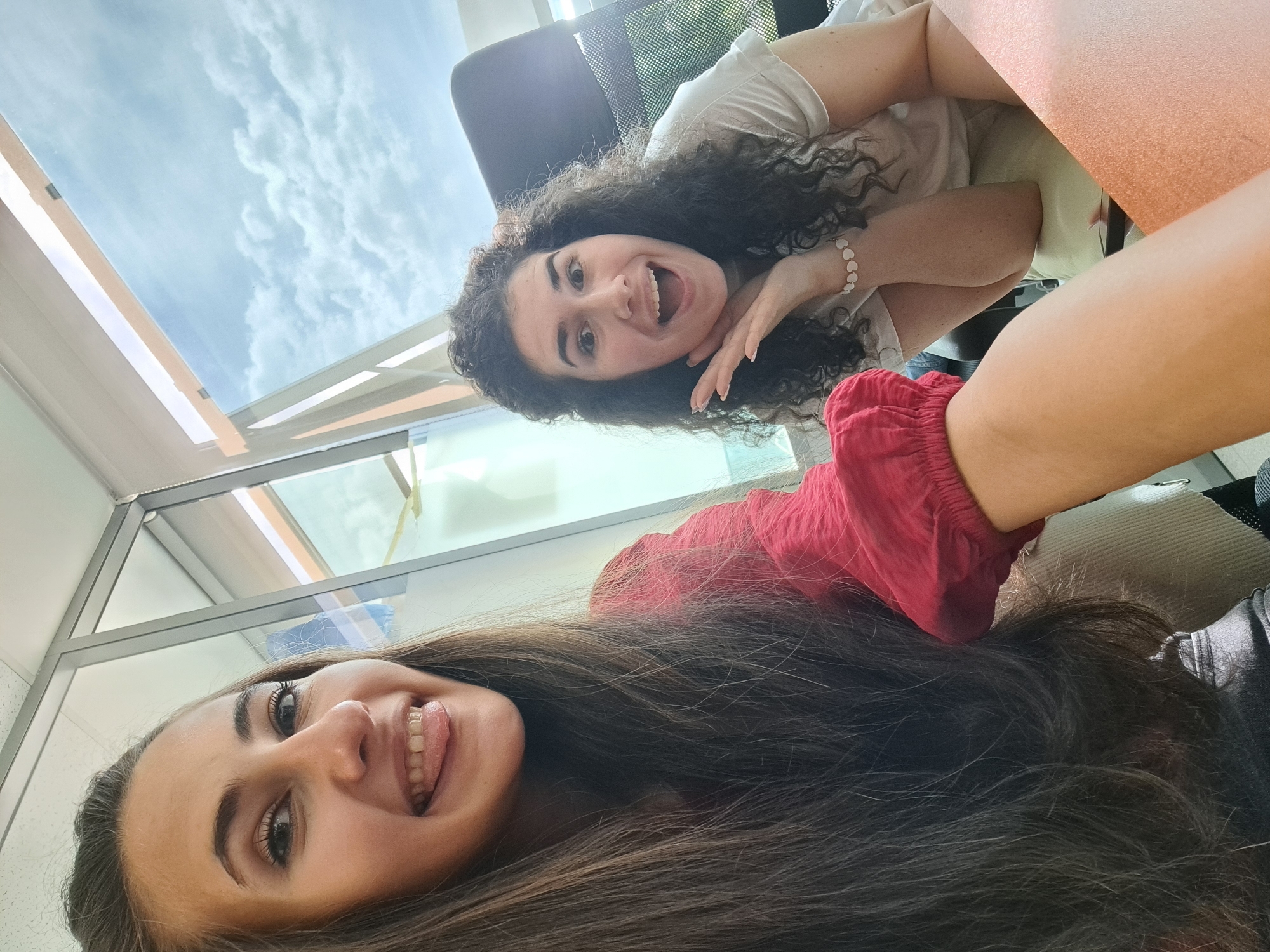 Two women are visible in the image. One has long, brown hair and is wearing a red shirt. She has her tongue slightly visible between her teeth and is smiling. The other woman has curly dark hair, a light-colored shirt, and a bracelet. She is smiling with her mouth open. They appear to be indoors, as there is a window in the background showing a cloudy sky. Based on the architectural style of the window and the indoor setting, it is possible they are located in a commercial or office building. Without specific architectural details or local landmarks, determining the exact location in Sofia, Bulgaria, is not possible.