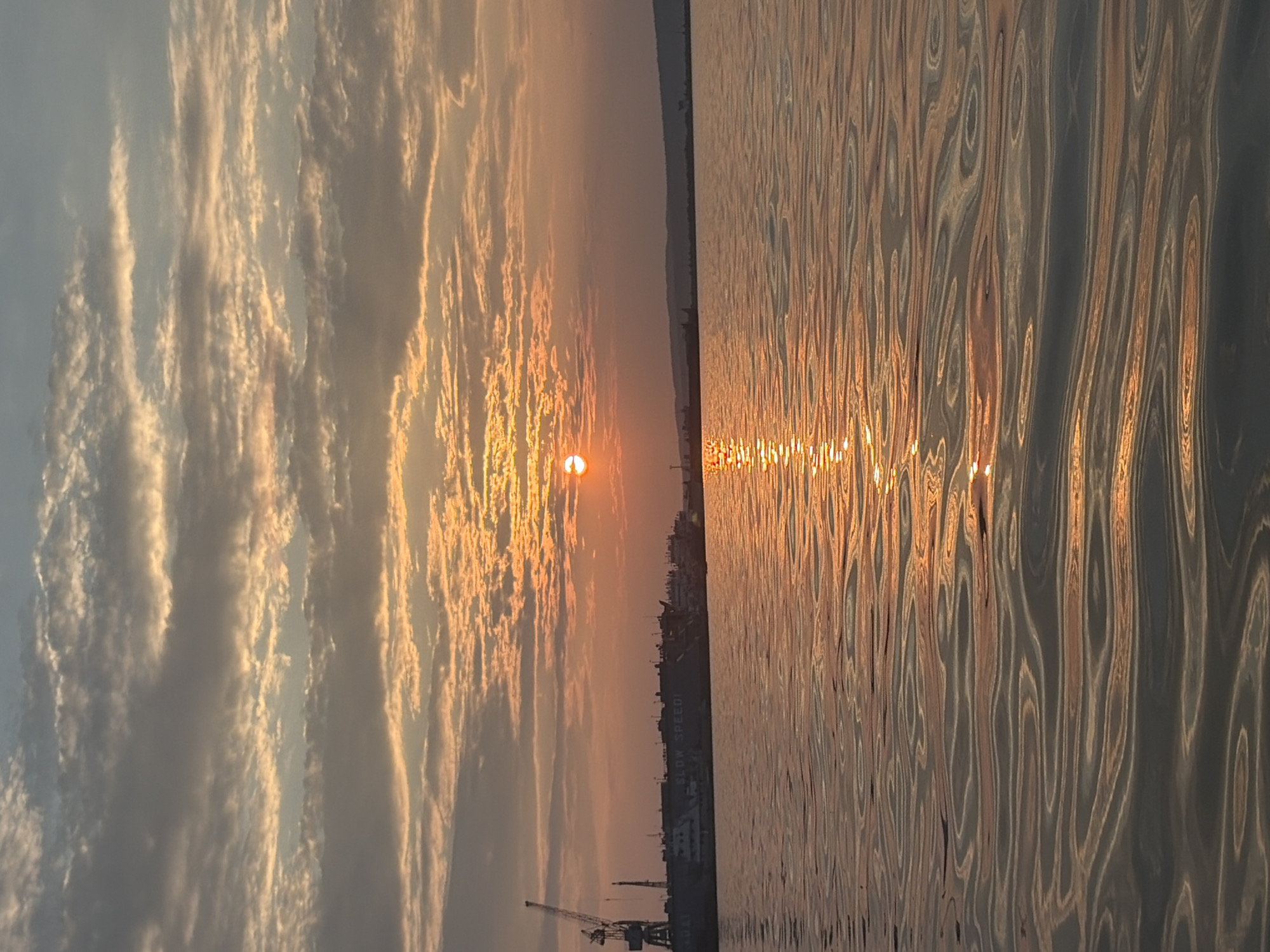 The image depicts a sunset over a body of water, likely the Kama River in Chaikovsky, Russia. The sky is filled with a mix of gray and orange clouds, illuminated by the setting sun which appears as a bright orange orb. The sun's reflection creates a shimmering path across the rippling water in the foreground. In the distance, a silhouetted industrial area with cranes and buildings lines the horizon. The overall scene is tranquil and atmospheric.