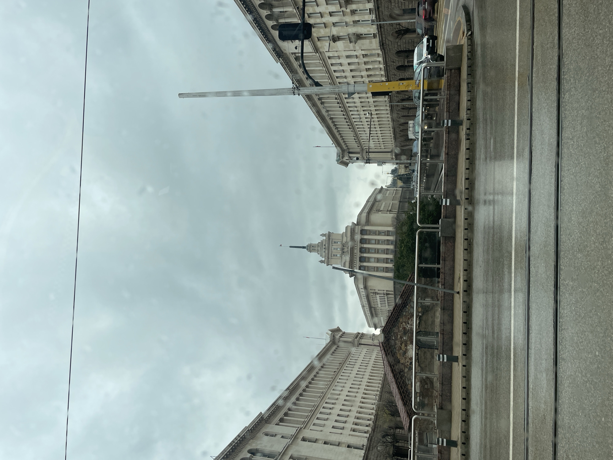 The image shows a view of buildings and street in Sofia, Bulgaria. The buildings are predominantly white with classical architectural details. In the center, there is a large building with a spire. The street is wet, possibly due to rain. There are trolleybus wires visible in the sky. The image appears to be taken from inside a vehicle looking up at the buildings. The street appears to have trolley tracks embedded in it. There are cars parked on the right side of the street.