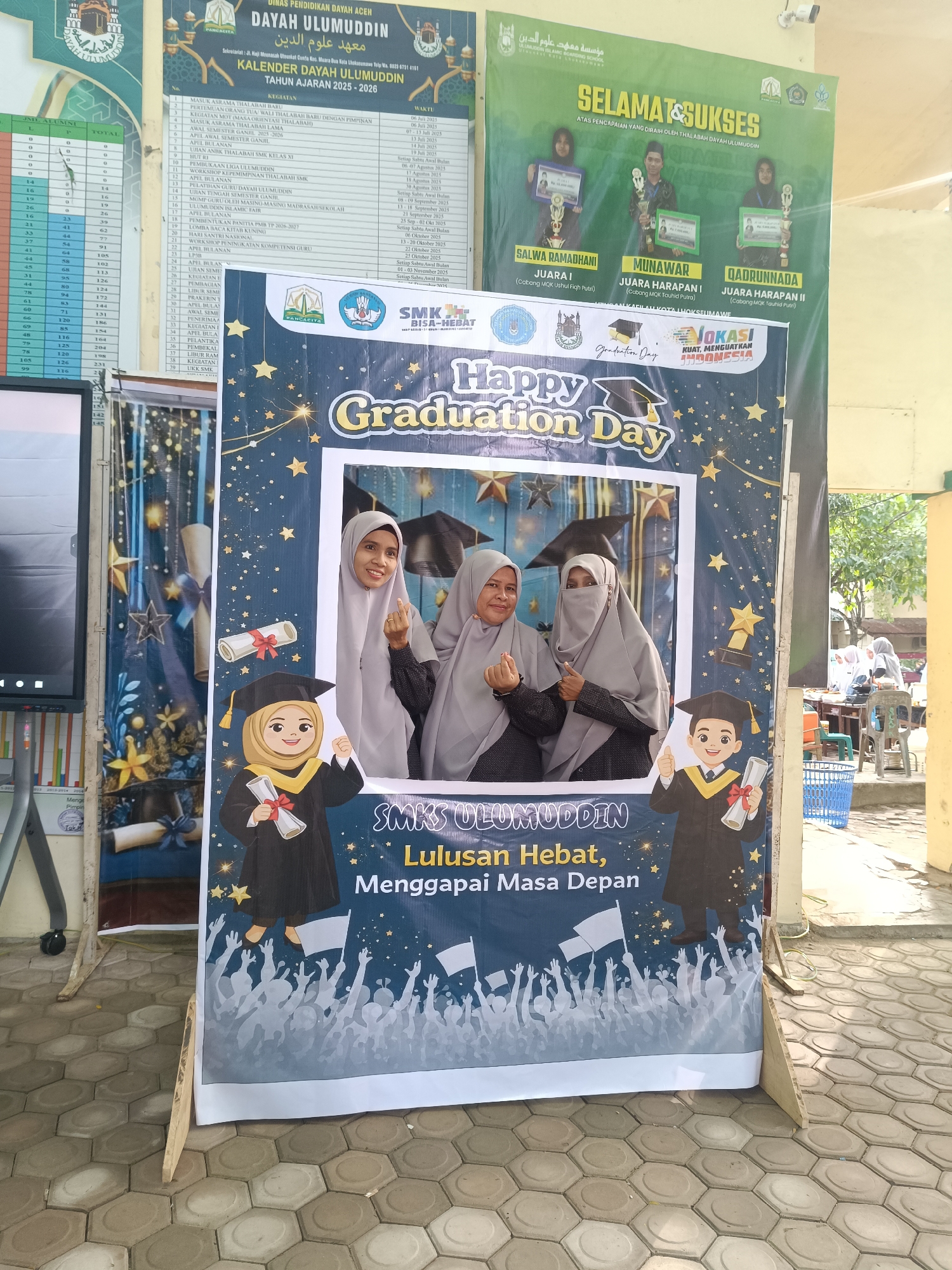 The image shows three women posing behind a "Happy Graduation Day" photo booth at SMK Ulumuddin, located in the city of Lhokseumawe, Indonesia. The booth is decorated with graduation-themed graphics, including images of graduates, stars, and graduation hats. Above the booth are two banners, one a school calendar for the academic year 2025-2026, and the other a "Congratulations" banner displaying photos of three students, Salwa Ramadhani, Munawar, and Qadrunnada, who have won awards in competitions (MQK Ushul Fiqh and Tauhid). The women are wearing hijabs, and two are making a hand gesture. The background shows the exterior of a building, possibly the school, with some people and objects visible in the distance. The flooring is tiled.