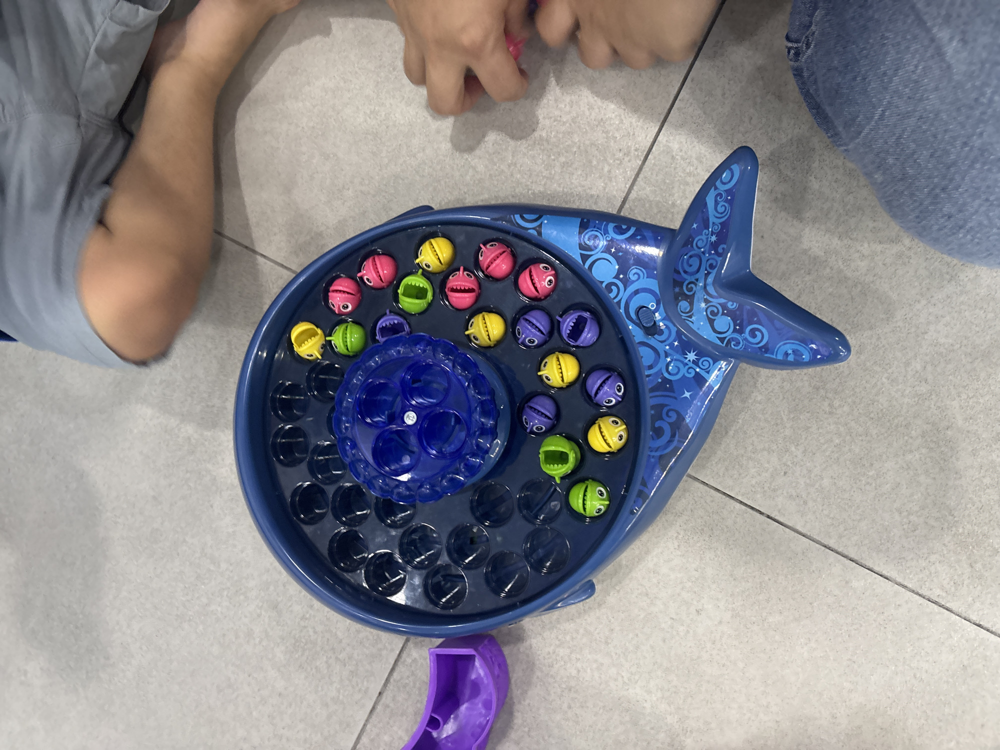 The image shows a blue whale-shaped fishing game on a tiled floor. The game has a circular spinning board with multiple holes, and colorful magnetic fish are placed inside. A purple ramp accessory is attached to the side of the whale. Two children's legs are visible in the background, suggesting they are playing the game. The lighting appears to be natural or from overhead, casting soft shadows. The scene is indoors, with the tiled floor suggesting a domestic or commercial setting. There is no information to indicate the location in Dubai, United Arab Emirates.