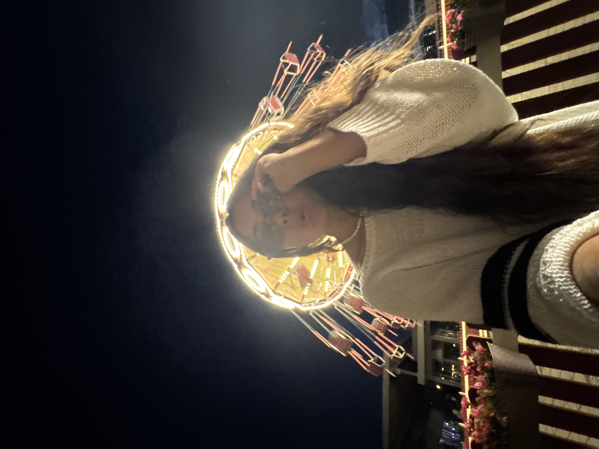 A young woman with long, dark hair is pictured at night. She is wearing a cream-colored sweater with black stripes on the sleeves and a pearl necklace. She is also wearing sunglasses and is holding them up to adjust them. Behind her, a brightly lit amusement park ride, possibly a swing ride, is in motion against a dark night sky. The ride has many illuminated bulbs and hanging seats. In the foreground, there are planters filled with pink flowers and striped seating. The scene suggests the woman is at an amusement park or fairground in Kazan, Russia.