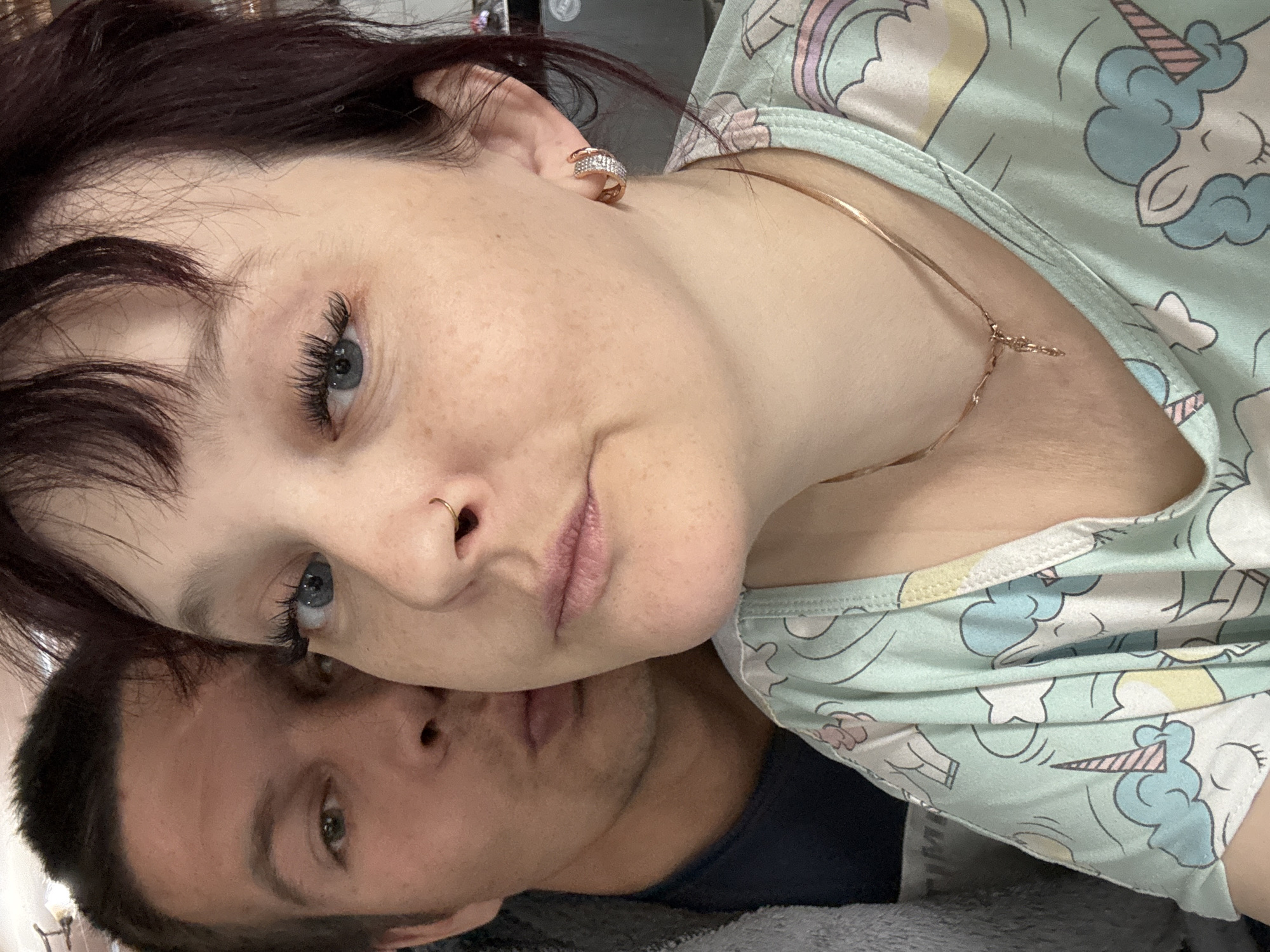 A close-up image shows two individuals. A fair-skinned woman with dark hair is positioned on top. She has blue eyes, freckles, and is wearing a gold earring and necklace. Her top is light blue with a unicorn pattern. Beneath her, a fair-skinned man with short dark hair is visible. He has green eyes and is wearing a dark blue shirt. The background appears to be a gray blanket. Location: Unknown, Unknown.