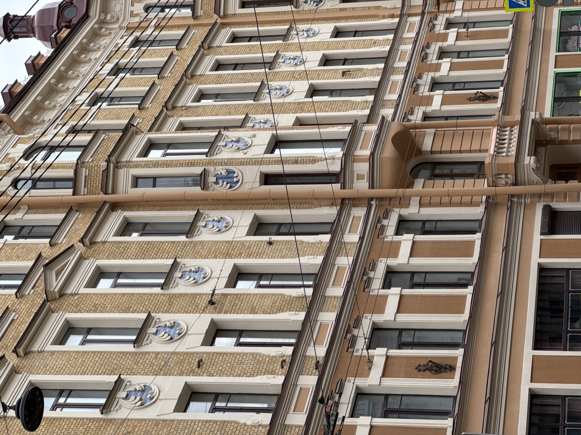The image shows a multi-story building in Saint Petersburg, Russia. The facade is detailed with light tan brickwork and decorative elements, including rounded blue-and-white ornamentation above the windows. Dark horizontal windows line each story. Electrical wires run diagonally across the facade. A light tan-colored vertical section with dark-framed windows is present on the right side of the building. A clock and a street sign are visible on the left side.