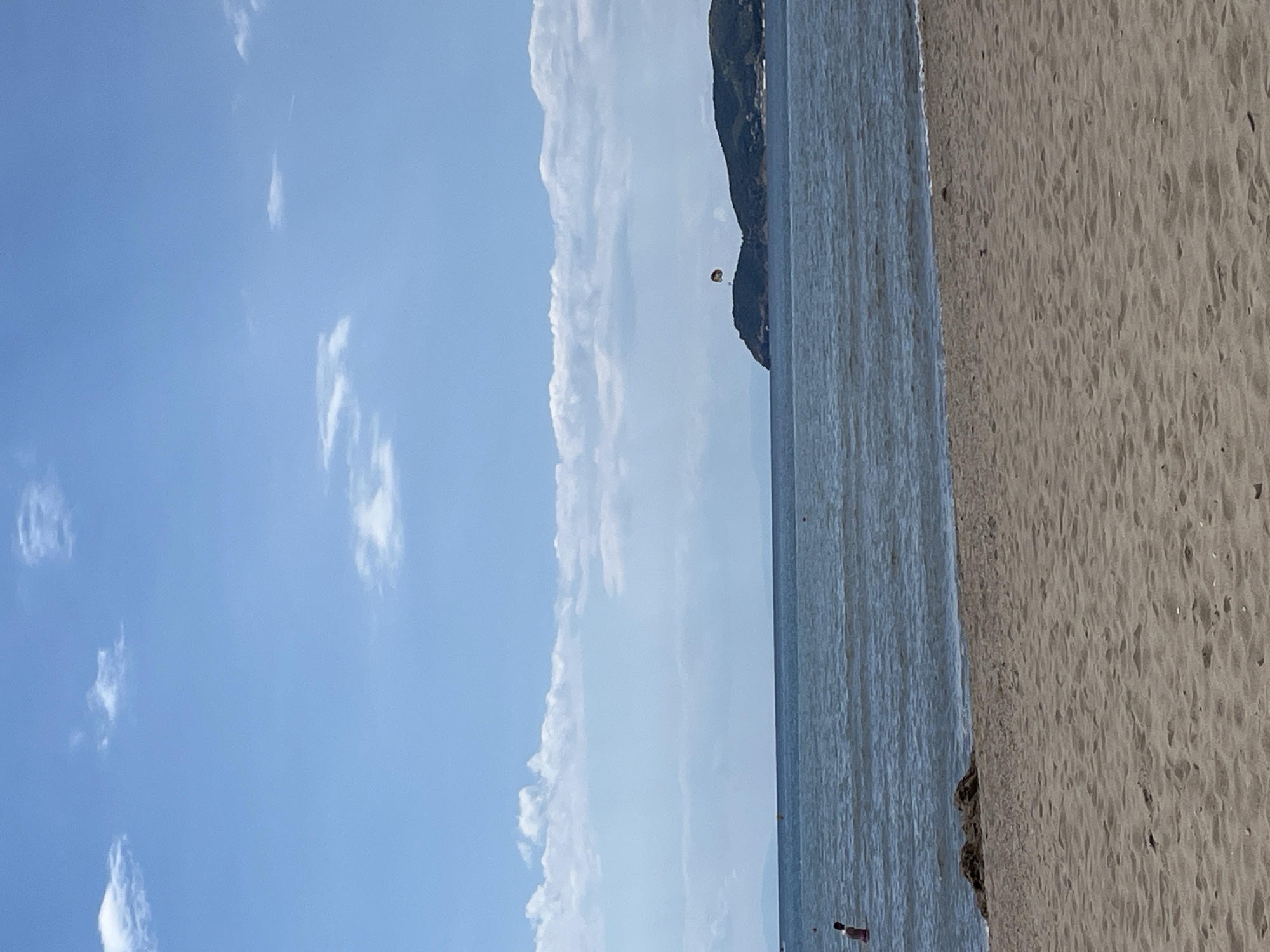 This is a photograph taken from a sandy beach looking out at the sea. The sky is blue with scattered white clouds. In the distance, there is a body of water with a visible horizon and a hilly landmass. A person is standing in the water on the left side of the frame. In the middle-right distance, a parasail can be seen in the air above the water, likely pulled by a boat out of frame. The foreground is dominated by the sand of the beach. The overall scene depicts a daytime outdoor recreational setting, likely a tourist location. The location is identified as Unknown, Greece.