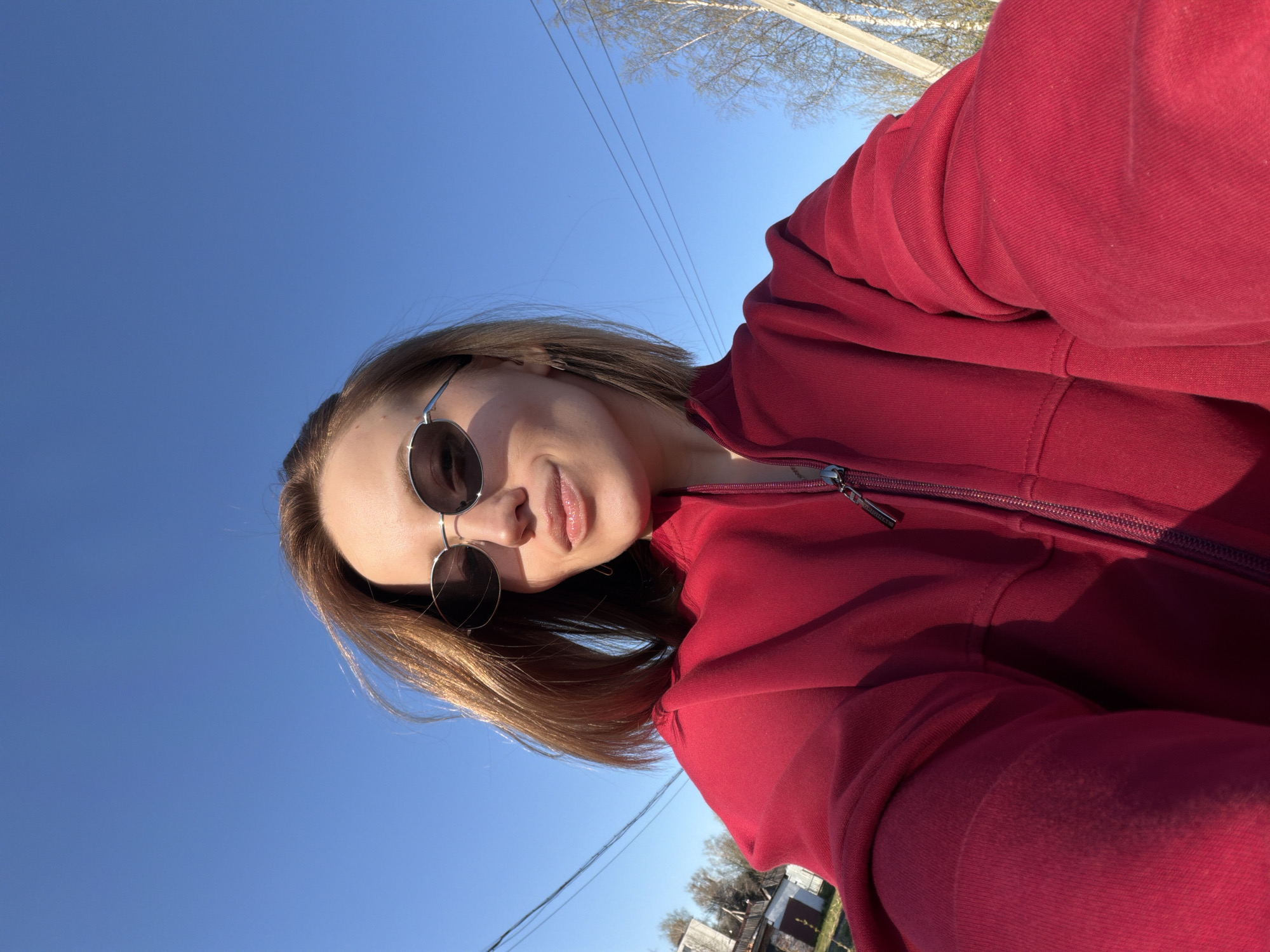 A woman with shoulder-length brown hair is wearing sunglasses and a red zip-up hoodie. She is outdoors under a clear blue sky. Power lines are visible in the background. In the distance, there are trees, and what appears to be houses. The location is Reshetnikovo, Russia.