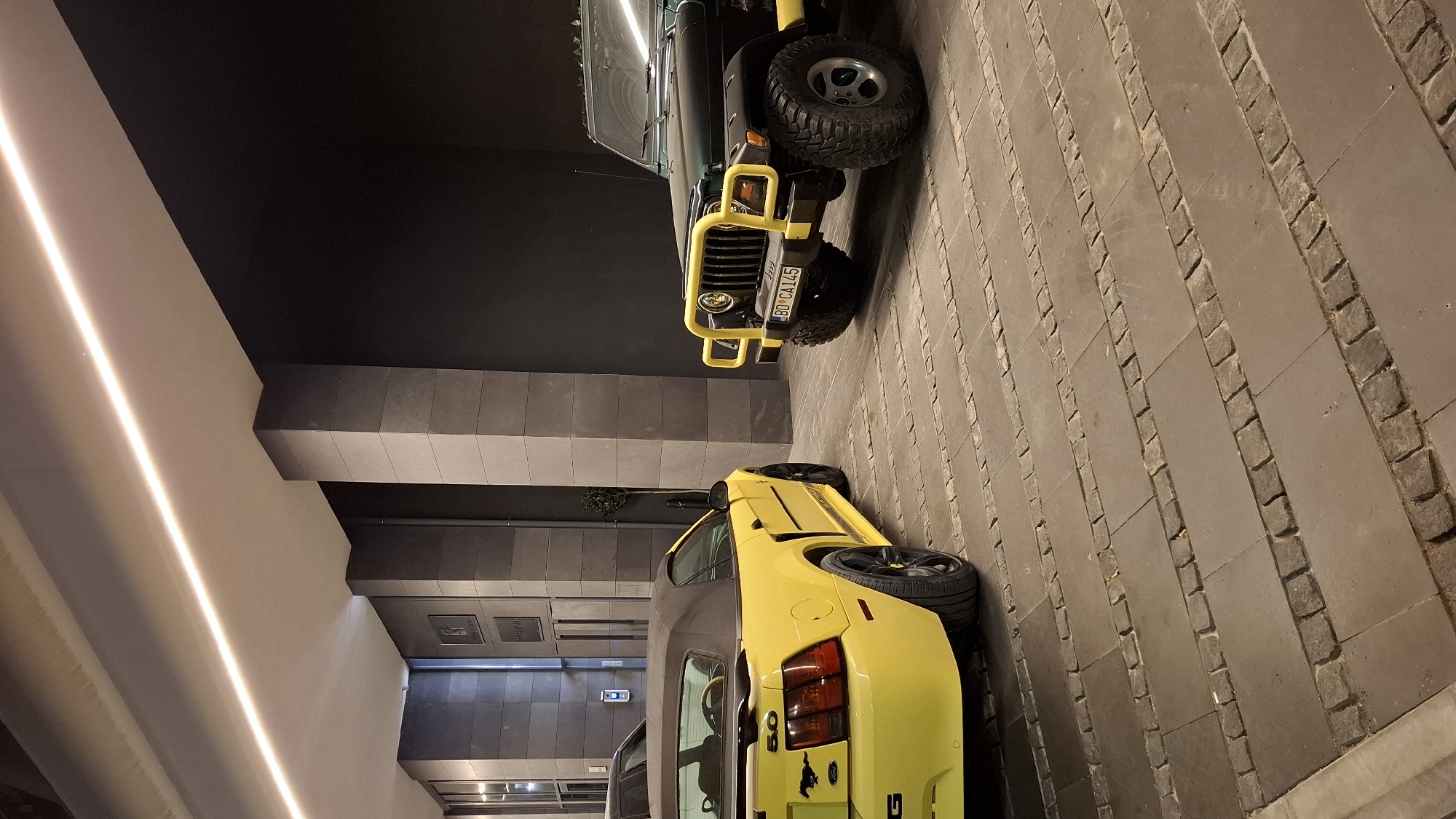 The image shows two vehicles, a yellow Mustang convertible and a dark-colored Jeep, parked on a paved surface. The paving consists of large rectangular stones with bands of smaller cobblestones. The vehicles are in a semi-enclosed space with a dark ceiling. The building architecture features recessed lighting and tiled walls. The license plate on the Jeep indicates the vehicle is from Budva, Montenegro.