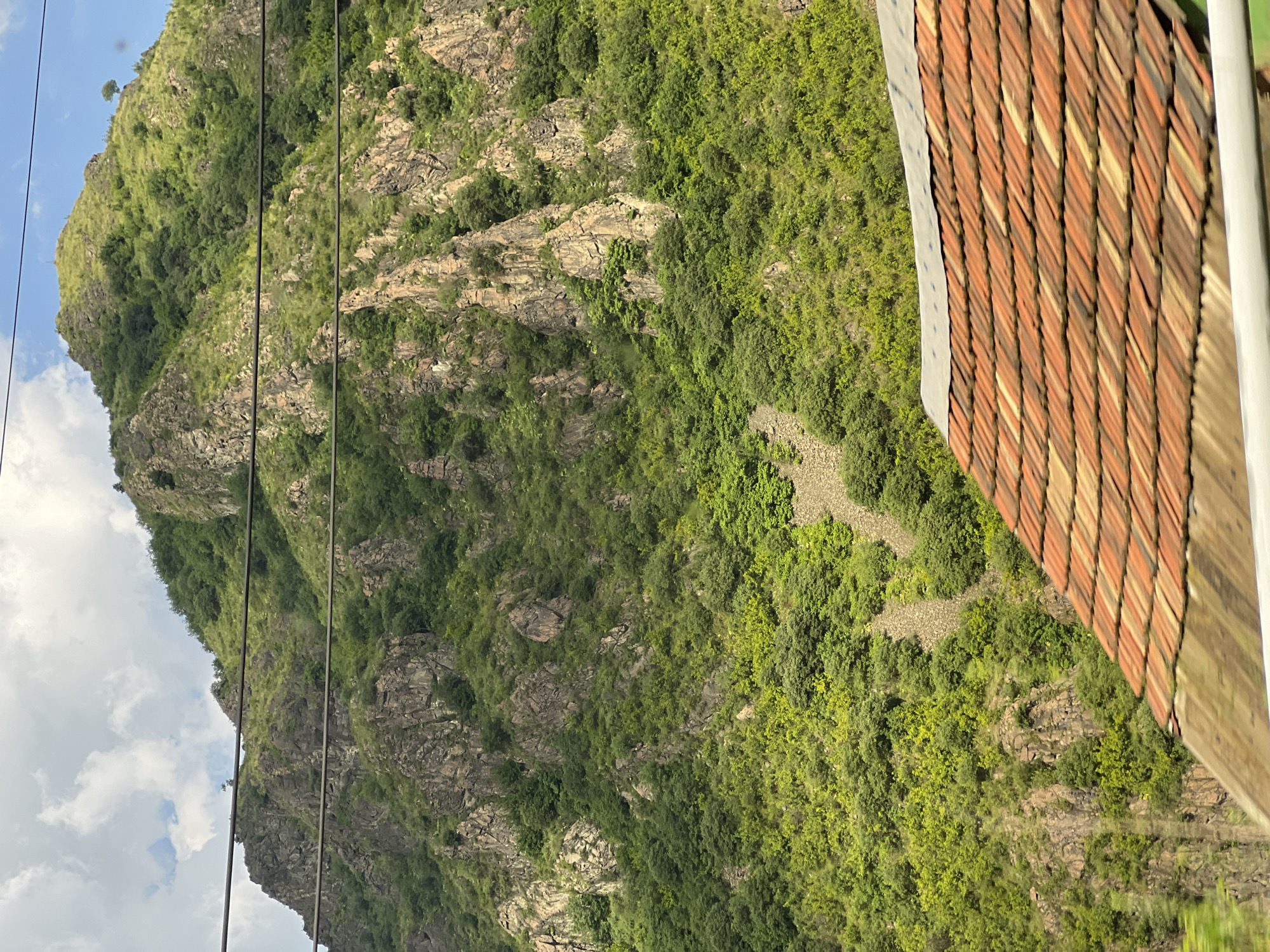 The image depicts a steep, rocky mountain slope covered in lush green vegetation and trees. In the foreground, the edge of a terracotta tiled roof is visible. Several dark power lines traverse the frame horizontally across the mountain. The sky above is partly cloudy with patches of blue. The overall scene suggests a natural landscape with some man-made elements, likely on the outskirts of a populated area. The mountain appears to be part of the landscape near Alaverdi, Armenia, a city known for its mountainous surroundings and industrial history, which might explain the presence of power lines.