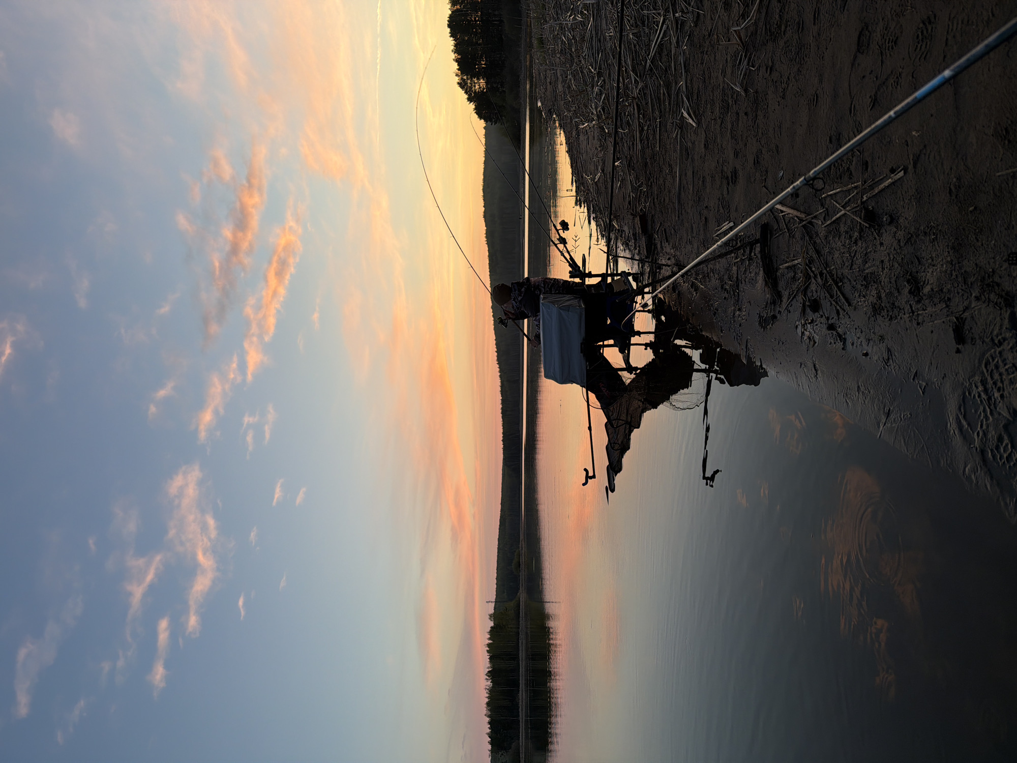 A person is fishing at sunset on the shoreline of what appears to be a body of water, possibly a lake or a reservoir. The location is likely in the city of Chaykovsky, Russia. The fisherman is seated with fishing rods extended into the water. The sky displays a gradient of colors from blue to orange as the sun sets. The opposite shore is lined with trees that are silhouetted against the light. The shoreline is muddy and has some reeds or plants. A fishing net and other equipment are near the person. The water surface reflects the colors of the sunset.