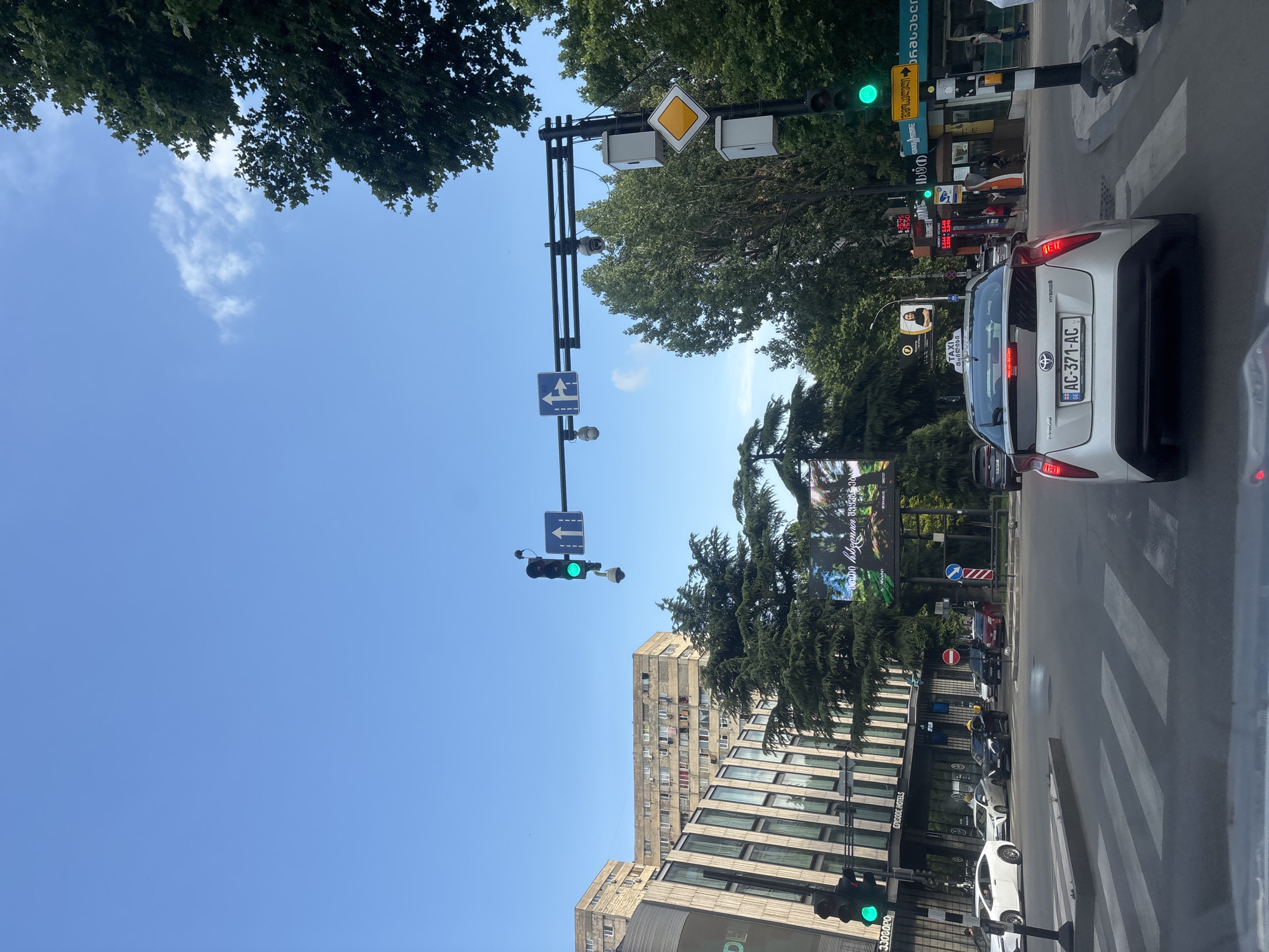 This image captures a street scene in Tbilisi, Georgia, on a clear, sunny day. The perspective is from inside a vehicle, looking forward and slightly upwards. The main subjects are the street, traffic signals, cars, and buildings.
In the foreground, a white Toyota Prius with a license plate reading "AC-371-AC" is visible. It appears to be stopped at a crosswalk. Several other cars are also present in the traffic, including a black car behind the Prius and some partially visible vehicles further down the street. A pedestrian crosswalk is prominent in the lower right of the frame.
Traffic lights are visible, with at least one showing a green light, indicating that traffic is moving or about to move. Several road signs are also present, including directional signs and what appears to be a warning sign. Some of these signs have text in Georgian.
The background features a mix of elements. Tall, modern buildings with numerous windows line the street, one of which has "EPISODE HOTELS" signage. Lush green trees are abundant, creating a canopy over parts of the street and softening the urban landscape. There's also a billboard visible in the distance with an image of a person.
The overall scene suggests a typical urban environment in motion, likely during daylight hours given the bright sunshine and clear blue sky. The presence of multiple cars and traffic control devices indicates a busy intersection. No people are clearly discernible as the primary subjects, though some figures can be seen in the background near shops. The text "TAXI" is visible on a sign, along with Georgian script.