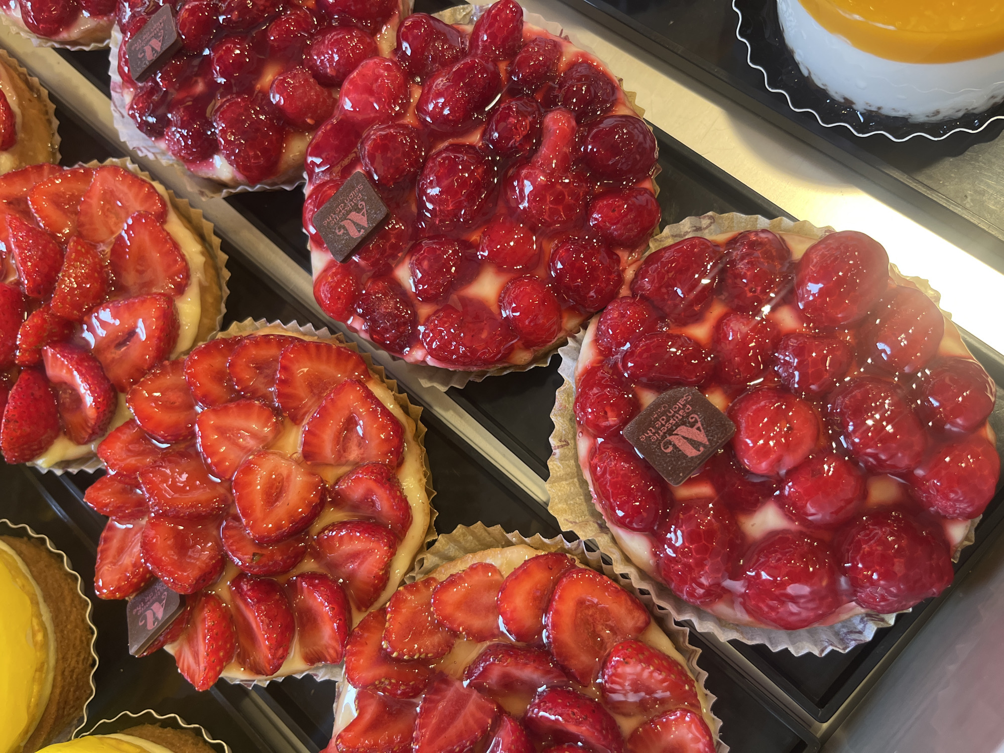 The image shows a close-up, overhead view of several fruit tarts displayed in what appears to be a bakery or pastry shop. The tarts are arranged in rows and are primarily topped with fresh strawberries and raspberries. Each tart has a golden-brown crust and a creamy, pale filling, possibly custard or pastry cream. Many of the fruits are glazed, giving them a shiny appearance. Some tarts feature small, dark chocolate squares with embossed lettering, which are likely decorative branding elements.
In the upper left corner, a portion of a yellow tart, possibly lemon or passionfruit, is visible. To the right of the strawberry tarts, a tart with raspberries is prominently displayed. In the bottom right corner, a circular dessert with a white base and a bright orange topping, likely a fruit coulis or glaze, is partially visible. The tarts are presented on a dark surface, possibly a display case. The overall impression is one of fresh, attractive pastries. The context provided identifies the location as a patisserie in Barr, France.
