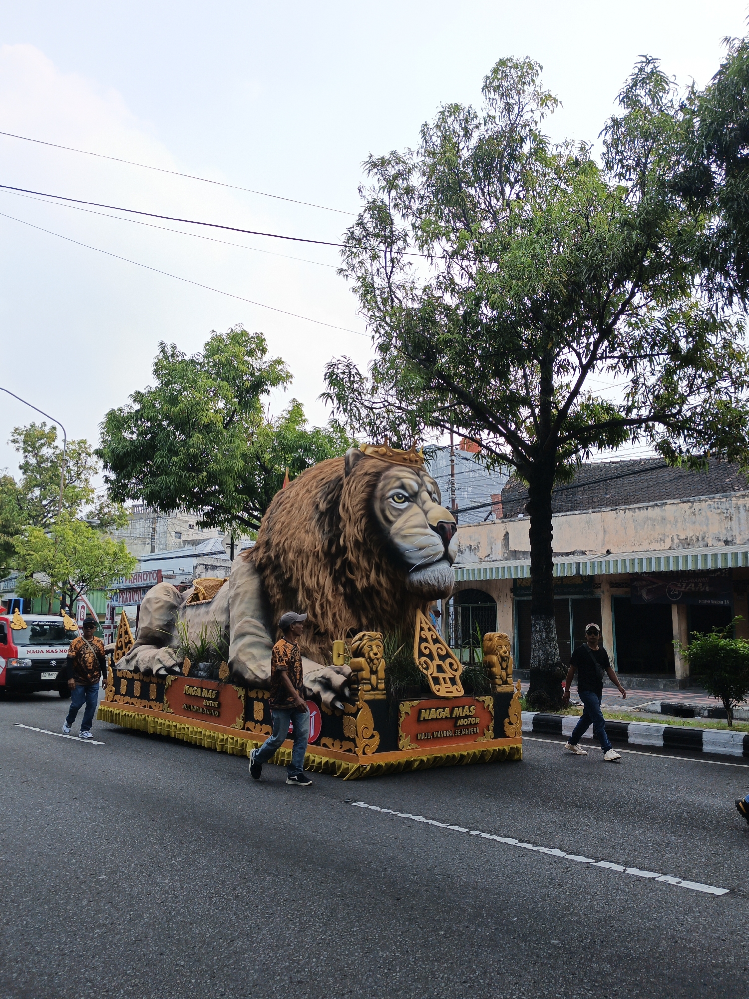 In Bareng Lor, Indonesia, a parade float featuring a large, detailed sculpture of a reclining lion with a golden crown is moving down a street. The float is decorated with golden ornamental elements and the words "NAGA MAS MOTOR MAJU, MANDIRI, SEJAHTERA". Two men in matching orange shirts with lion motifs walk alongside the float. Further down the street, another man in a black shirt and blue jeans walks across the road. A red truck with the words "NAGA MAS MO" is visible on the left. The street is paved and has white lane markings. Trees line the background, with a building featuring awnings visible behind them. The sky is overcast.