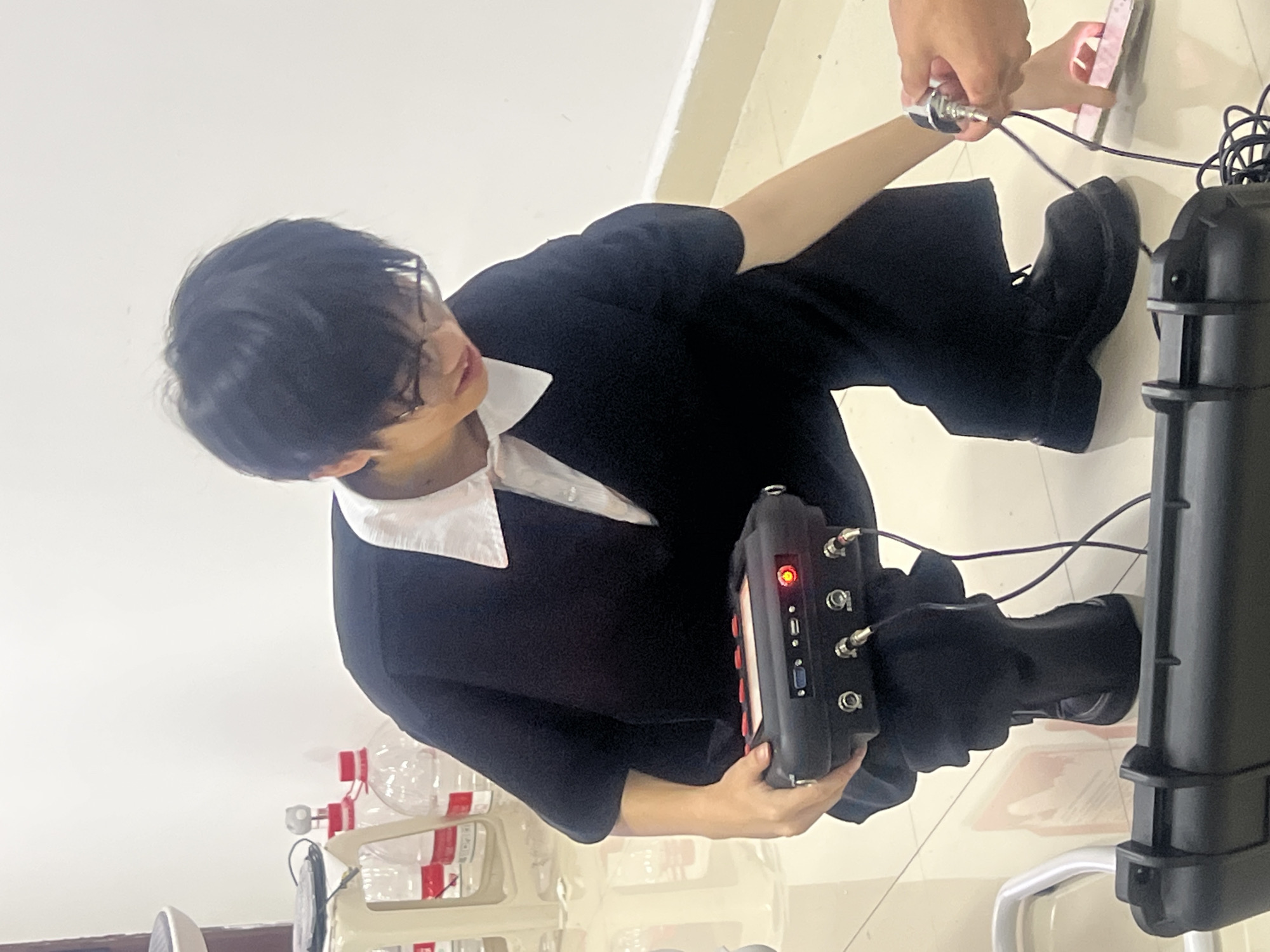 An individual with dark hair, glasses, and wearing a white collared shirt under a black top and black pants, sits on the floor, holding a black device with multiple inputs and a red light. Various cables connect the device to a larger black case and another object being held by another individual, who is not fully visible. Several water bottles are visible in the background. The location is inside a building, possibly in Jiangjin, China, based on visual cues.