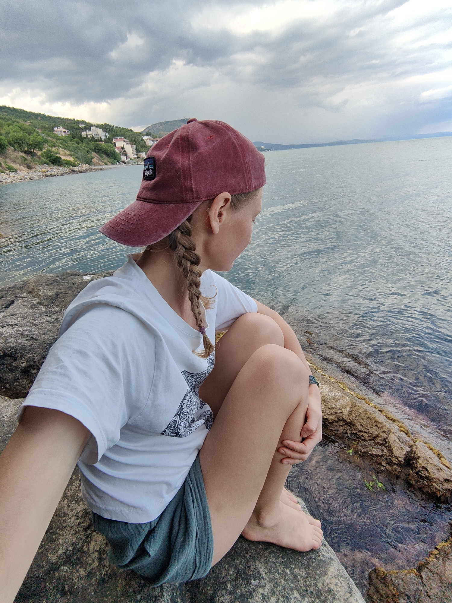 A young woman wearing a maroon baseball cap, white t-shirt, and green shorts sits on rocks by the sea. Her hair is in a braid. She is looking out at the water. The sky is cloudy and overcast. In the distance, there are hills with buildings on them. The location is Utios, Ukraine.