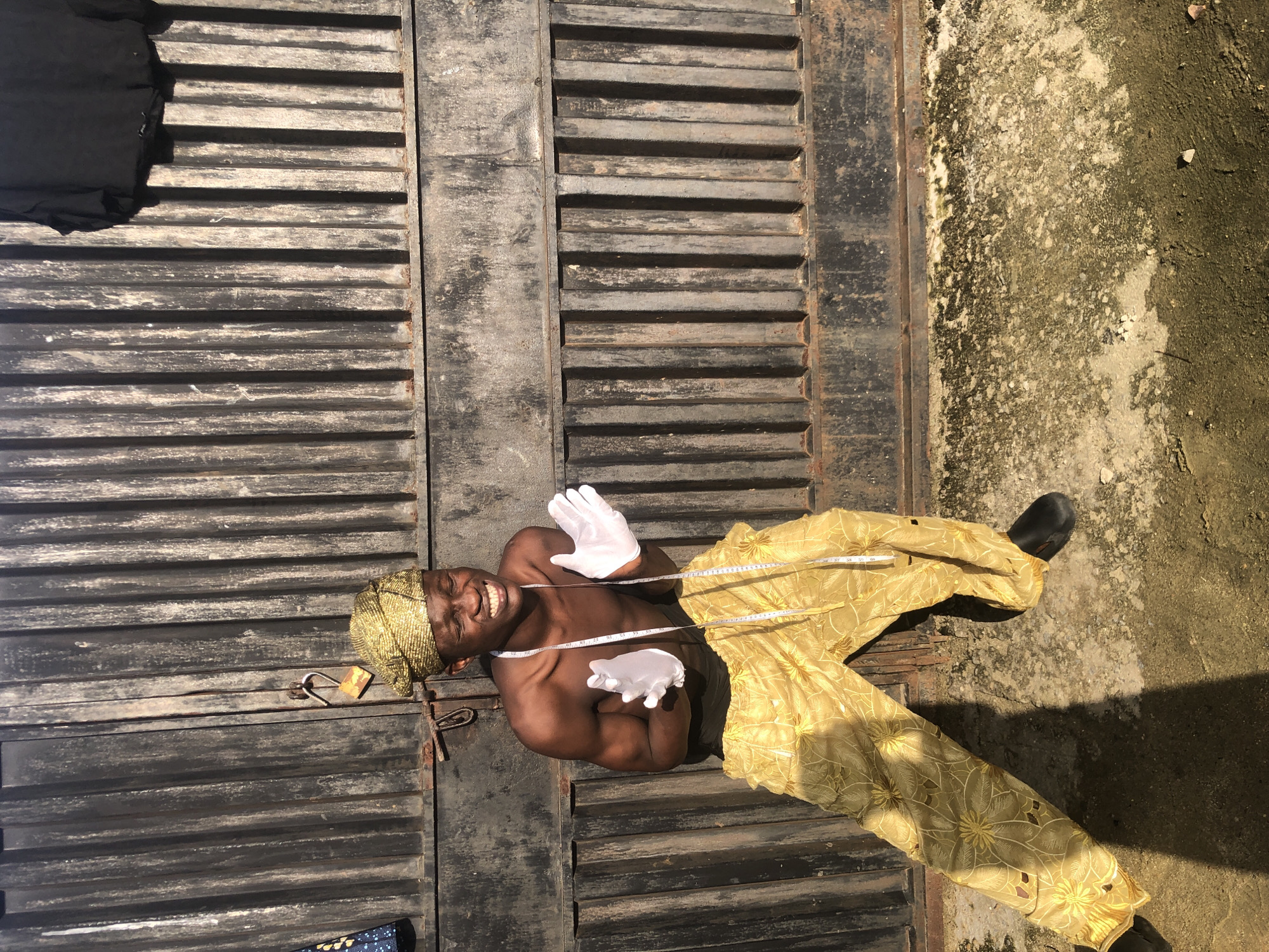 A young Black man, appearing to be in his late teens or early twenties, stands in front of a corrugated metal gate in Ado, Nigeria. He is shirtless, revealing muscular arms and chest. He wears a gold, textured headwrap and loose-fitting gold pants with a floral pattern. White gloves cover his hands, which are held out in front of him. A white measuring tape is draped around his neck. He has a wide, joyful expression, with his eyes closed and teeth bared. The gate appears old and weathered, with a padlock hanging open from a latch. The ground in front of the gate is a mixture of concrete and dirt, with some patches of moss or algae. Shadows are cast by the man and the gate, indicating bright sunlight. A black shirt is hanging from the top of the gate on the right side of the frame. The overall scene is outdoors, with the sun clearly visible.