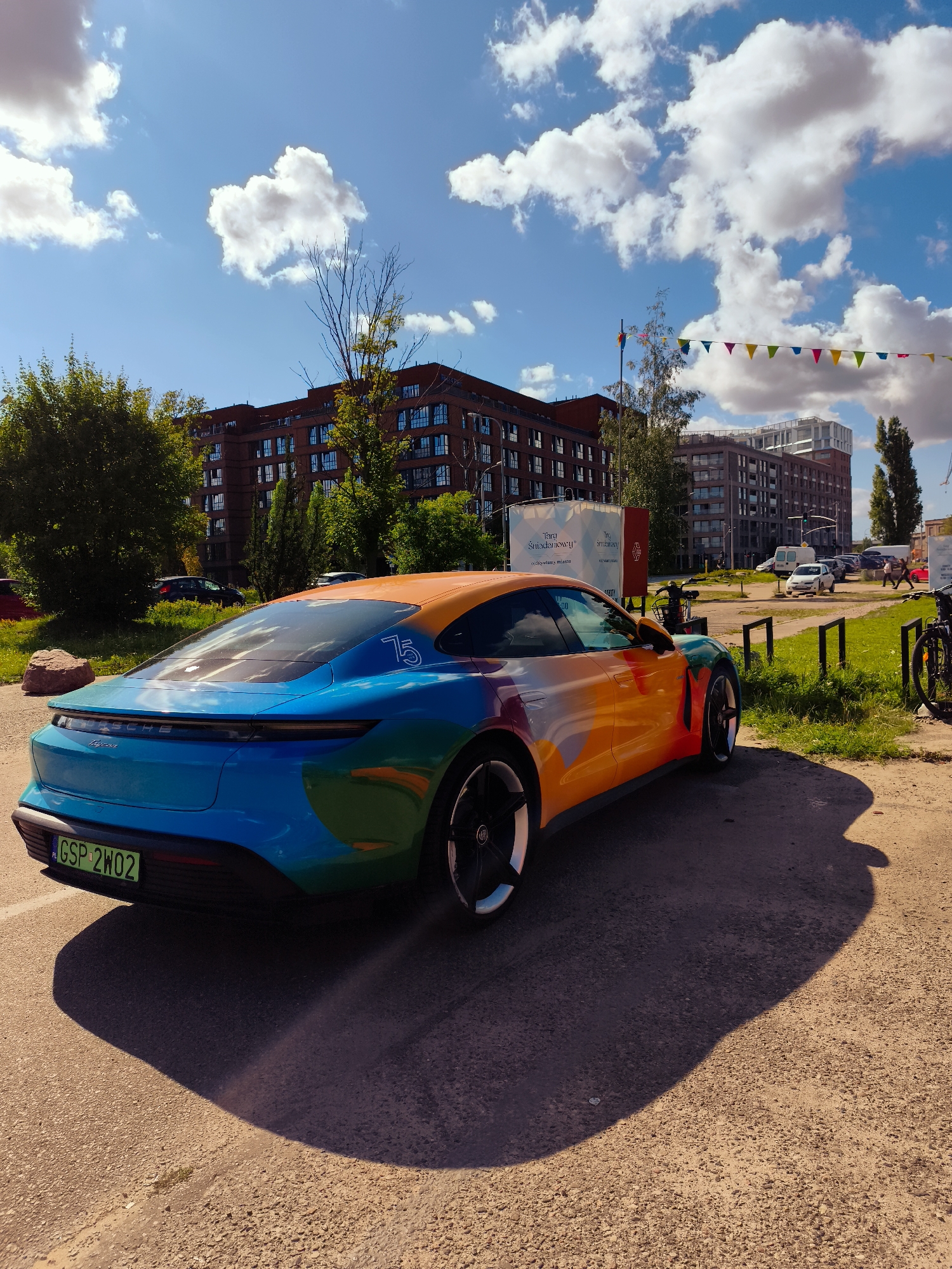 The image shows a vibrantly colored Porsche Taycan parked on an asphalt surface, casting a long shadow to its right. The car's exterior features a gradient of colors, transitioning from blue at the rear to orange at the top. The license plate reads "GSP 2WO2". In the background, several multi-story brick buildings are visible, some with numerous windows. Trees with green foliage are interspersed between the buildings. Overhead, a bright blue sky is dotted with fluffy white clouds, and a string of colorful triangular flags is strung across the scene. The environment appears to be an urban setting, possibly an event or exhibition space, given the festive flags and the presentation of the car. While the exact location in Gdańsk, Poland is not specified, the presence of Polish license plates and the architectural style of the buildings suggest it is within the country. There are no people clearly visible in the immediate vicinity of the car.