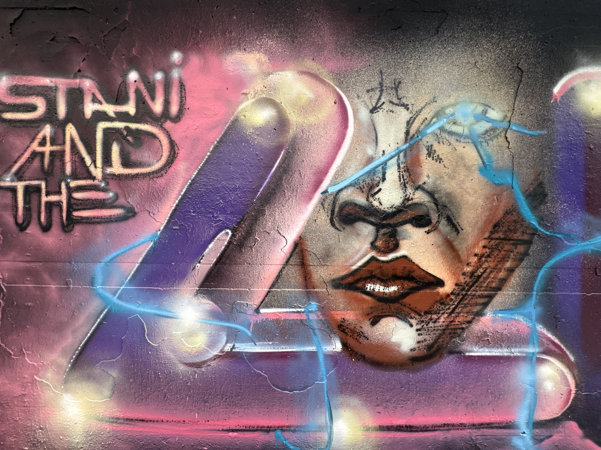 The image displays graffiti on a textured wall in Augsburg, Germany. The graffiti features bold, colorful lettering that appears to read "STANI AND THE 4". Superimposed over the text and a large, stylized number "4" is a portrait of a face with prominent lips and a nose. The color palette includes pinks, purples, blues, and browns, with spray paint effects creating a vibrant and dynamic composition. The wall itself shows signs of wear, with visible cracks and peeling paint, indicating it is an outdoor urban environment. The graffiti style is consistent with street art found in public spaces.