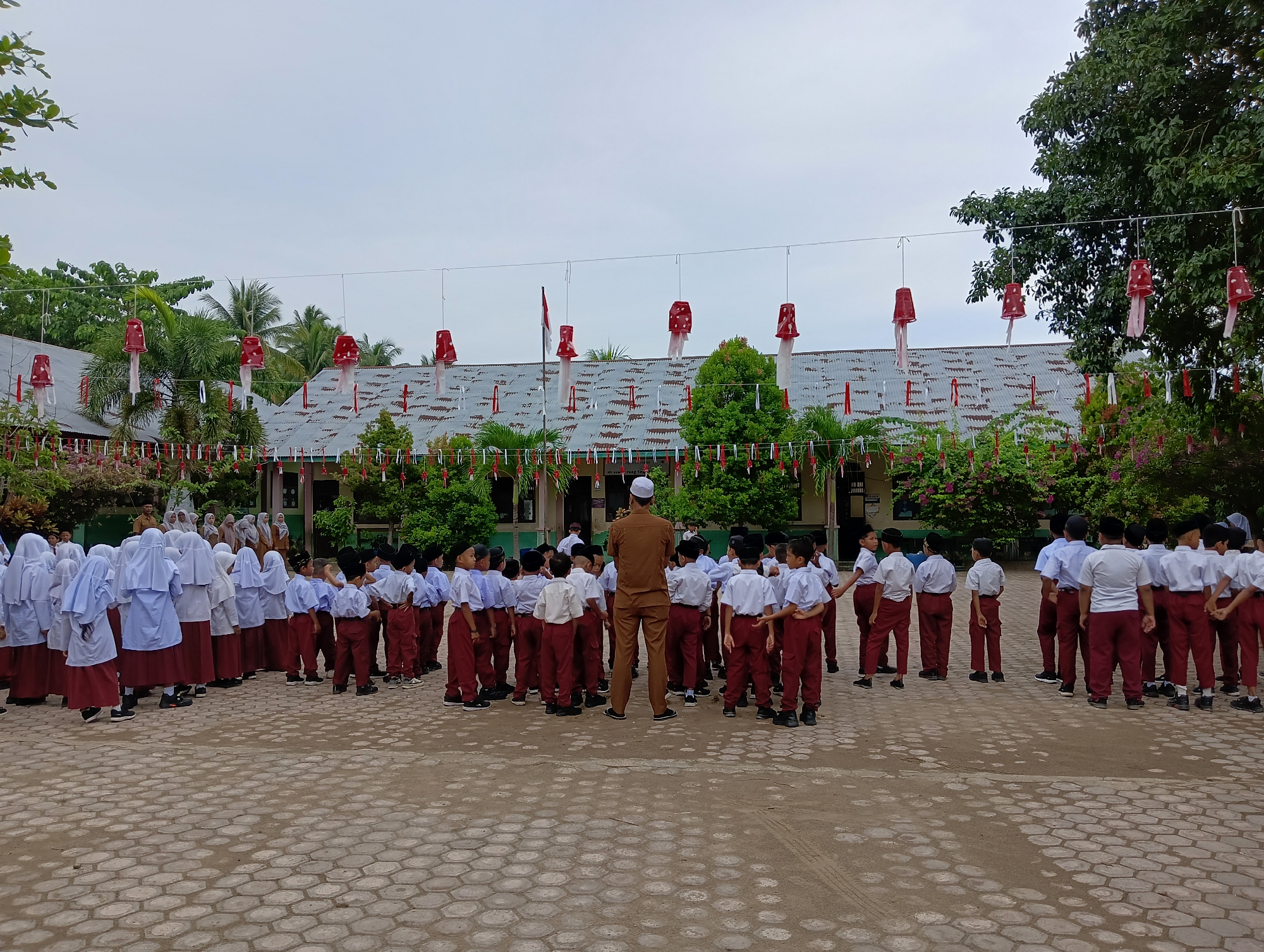 In the city of Baratpayaitek, Indonesia, a group of schoolchildren are gathered in an outdoor courtyard. The children, some wearing white hijab and uniforms, others in white shirts and maroon pants, are lined up facing a man in a brown uniform and white skullcap. The courtyard is paved with hexagonal interlocking bricks. In the background, a single-story building with a corrugated metal roof is visible, decorated with red and white banners and hanging red paper decorations. Trees and lush green foliage surround the building. The sky is overcast.
