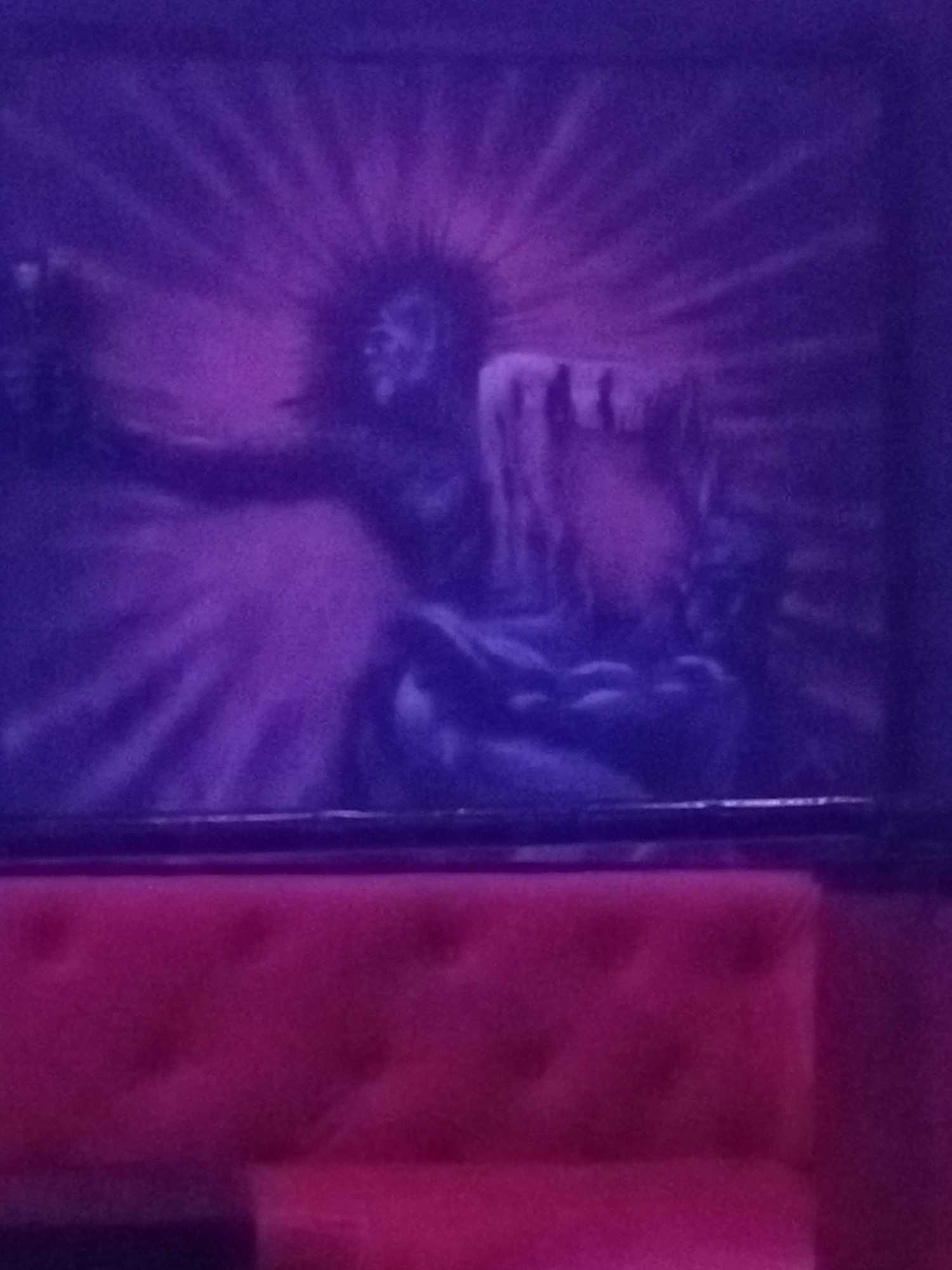 A dimly lit interior space, featuring a large framed painting above a pink tufted sofa. The painting is rendered in shades of blue and purple and depicts a seated figure with outstretched arms and radial lines emanating from their head. The environment is dark. The location is Unknown, Unknown.