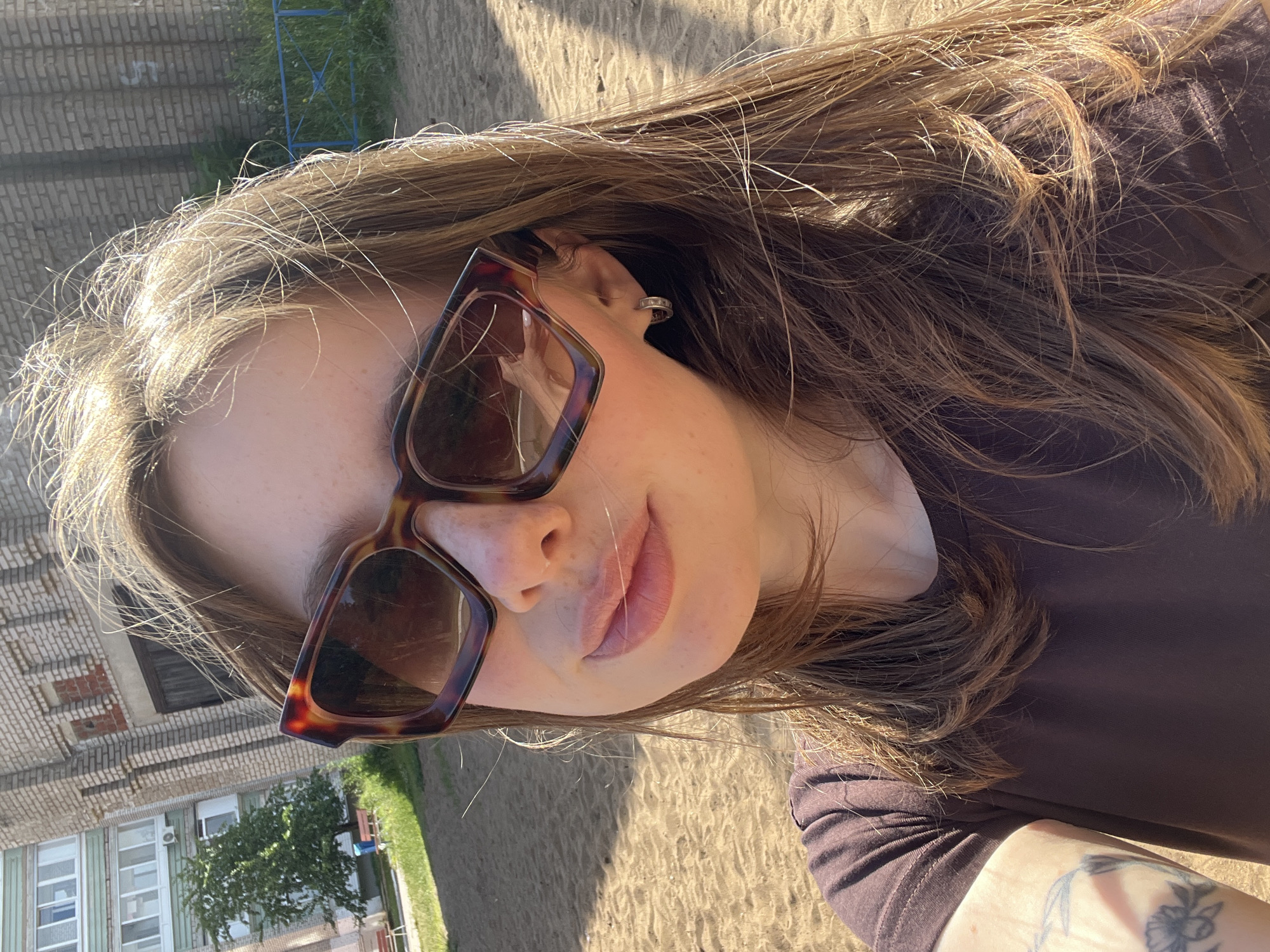 A close-up, rotated portrait of a young woman wearing large, tortoise-shell patterned sunglasses. She has long, brown hair that is styled loosely and falls around her face and shoulders. The woman is wearing a dark brown t-shirt, and a portion of a tattoo is visible on her left arm. Her lips are slightly parted, and she appears to be looking towards the camera. The lighting suggests it is daytime, with sunlight casting shadows on the sandy ground in the background, indicating an outdoor setting. In the blurred background, there are hints of buildings and greenery, consistent with a residential or park area. The image has a casual, candid feel.