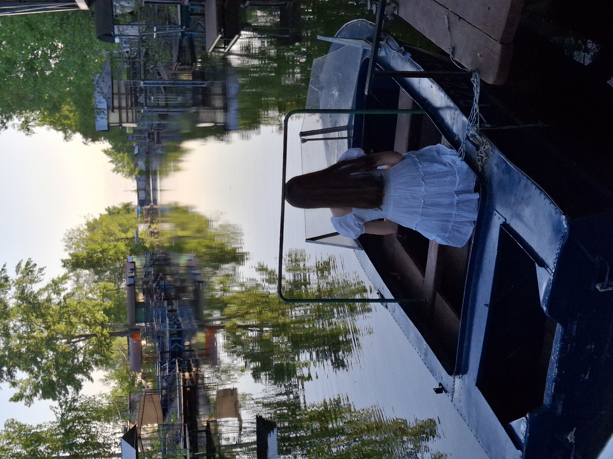 A young woman in a white dress sits in a blue boat on a river in Chaikovsky, Russia. The surrounding area features trees, buildings, and their reflections in the water. The boat is tied to a wooden dock.