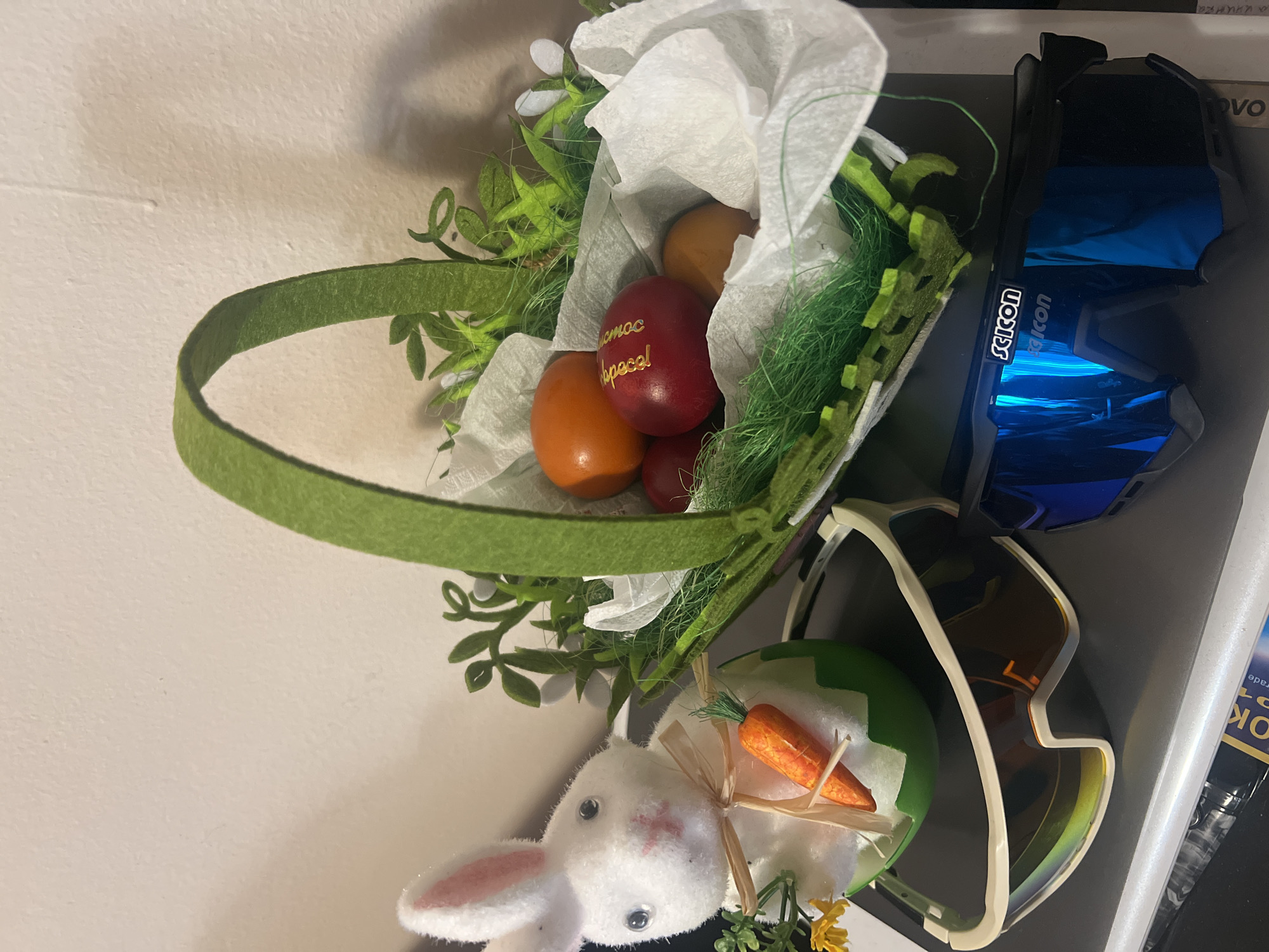 A close-up shot, rotated to be viewed sideways, presents an Easter basket with dyed eggs, accompanied by a white plush bunny and two pairs of sports sunglasses, possibly indoors. The basket, featuring a green felt handle, holds several eggs; one red egg has Cyrillic script written on it. The bunny, positioned near the basket, has a carrot tied to it. Adjacent to the basket are two pairs of sports sunglasses: one with black frames and reflective blue lenses, and another with white frames and brown lenses. The background is a pale wall. The location in Plovdiv, Bulgaria is unknown.
