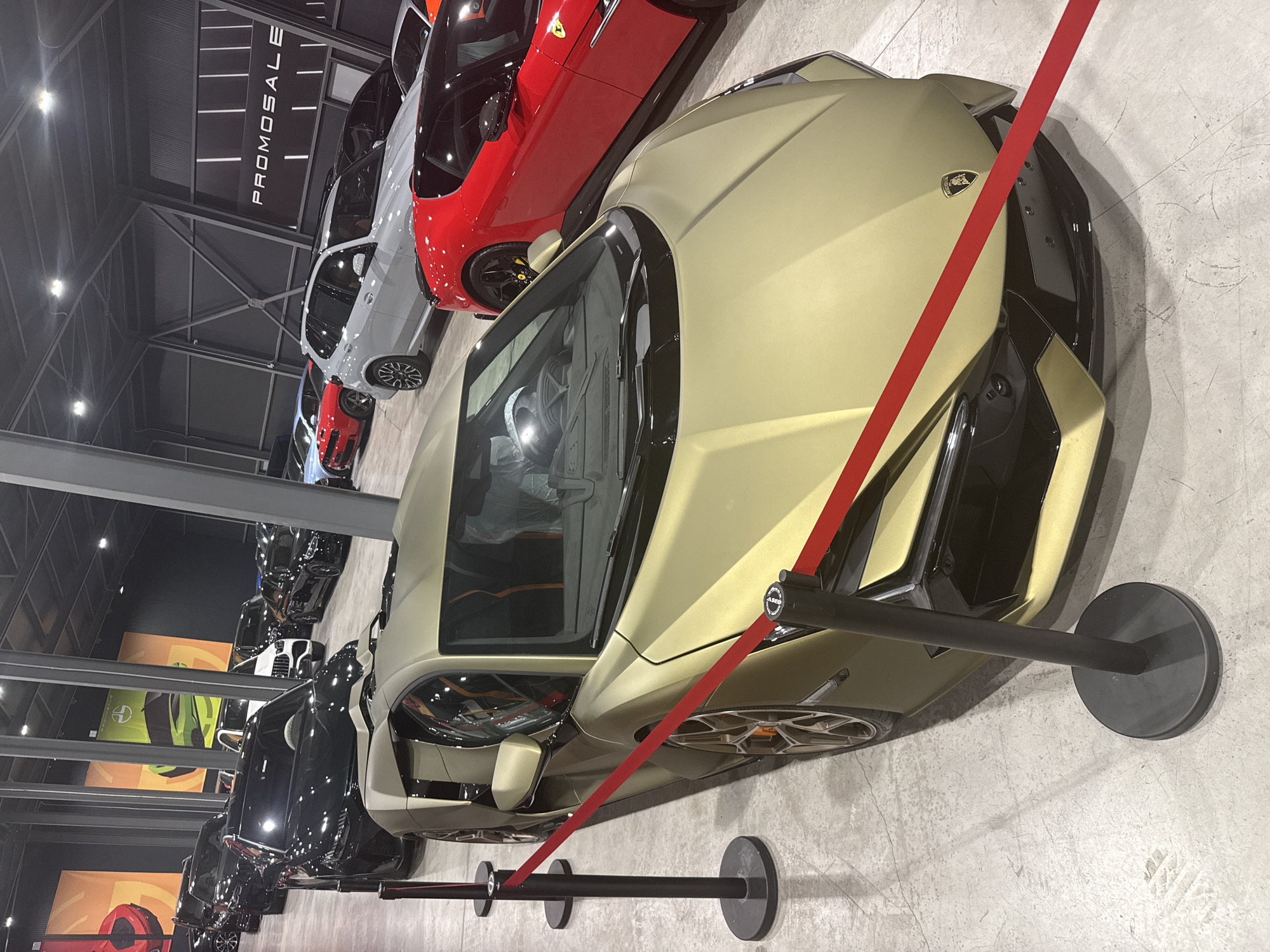 The image depicts the interior of a showroom, likely a car dealership, filled with a variety of vehicles. The primary focus is on a gold Lamborghini parked prominently in the foreground, with a red ribbon draped across its front. Several other cars are visible in the background, including black SUVs, a red sports car, and a light-colored SUV. The showroom features industrial-style lighting and exposed ceiling structures, characteristic of a modern commercial space. Banners displaying automotive imagery and branding are hung on the walls, with one banner visible on the right side displaying the text "PROMOSALE". The floor is concrete, with some tire marks visible. There are no people clearly visible in the image. The location is identified as Plovdiv, Bulgaria.