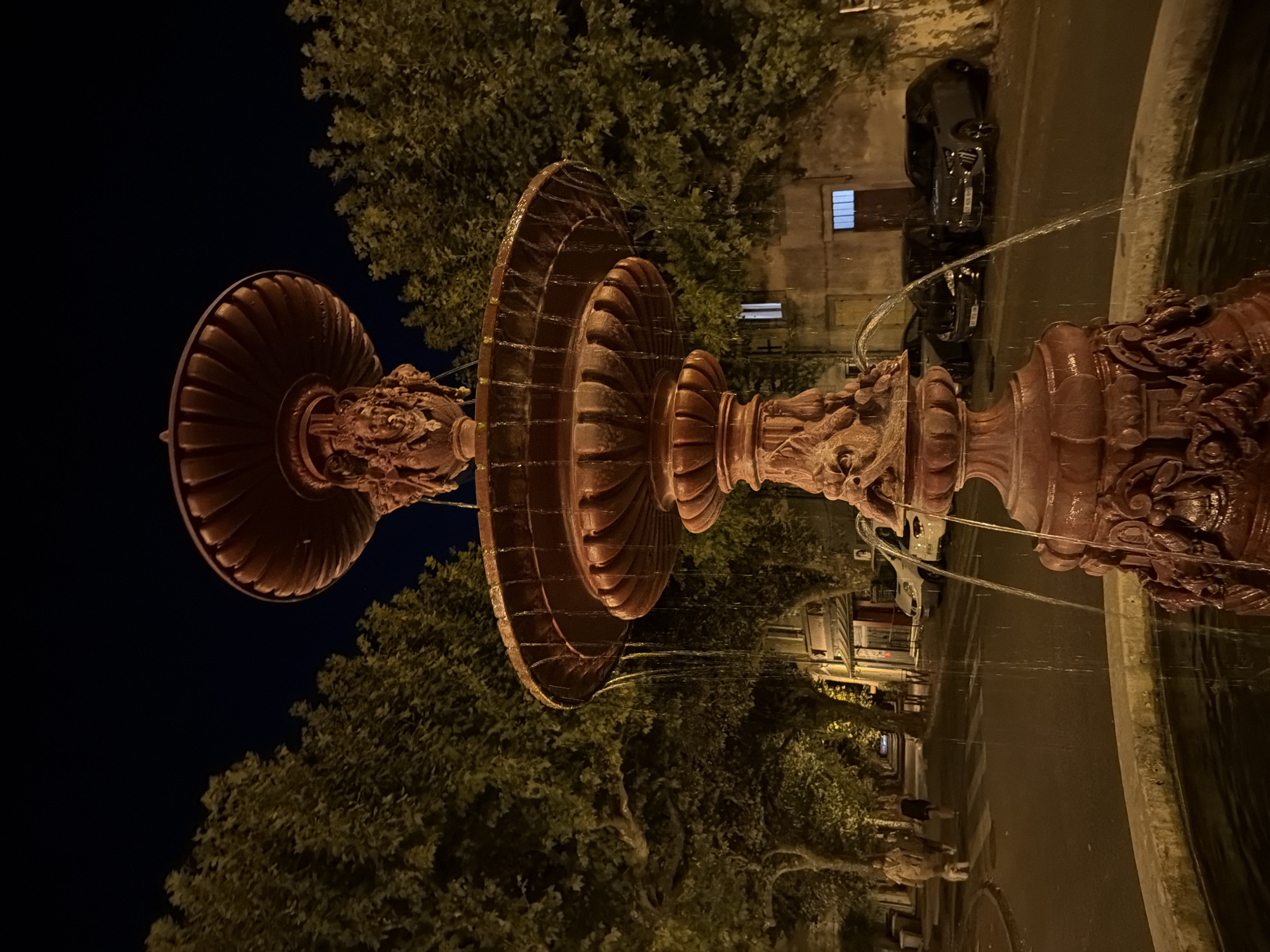 This is a nighttime, medium-distance shot of a red, ornate, multi-tiered fountain situated in a city square in Viviers, France. The fountain, made of stone or metal, features detailed carvings of faces and mythological figures. Water cascades down the tiers, catching the light from nearby streetlamps. In the background, a street is visible with parked cars, including a dark gray SUV and a black sedan. Buildings line the street, with illuminated windows suggesting occupancy. Trees with dense foliage flank the street, and the dark sky indicates late evening or night. Two figures, a woman and a man, walk a small white dog on the sidewalk to the left of the fountain. The overall atmosphere is calm and urban, with a touch of rustic charm from the fountain's design.