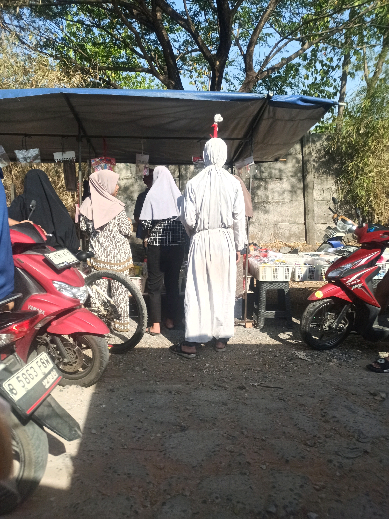 This image depicts an outdoor scene, likely a marketplace or roadside stall, in Telagamurni, Indonesia. The foreground features two red motorcycles. The motorcycle on the left has a license plate that reads "B 5863 F8H". Behind the motorcycles, a blue tarp covers a stall where several individuals are present. One person is wearing a long white robe and a white head covering. Another person to their left is wearing a beige hijab and a patterned dress. To the left of them, a person in a black hijab is partially visible. A person to the right of the white-robed individual is wearing a grey hijab and a checkered shirt. The stall displays various items in white plastic baskets. The background shows a concrete wall and dense vegetation, with trees overhead. The lighting suggests it is daytime.