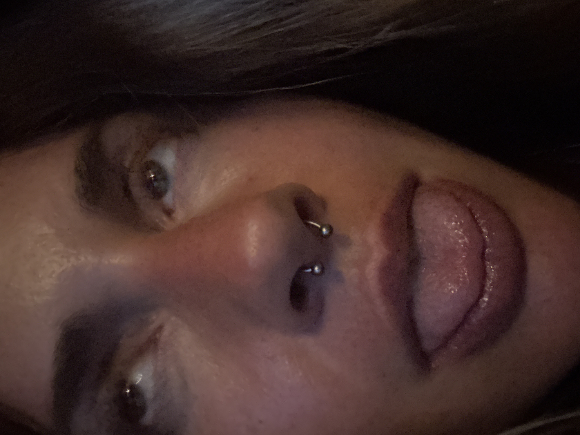 A close-up, indoor, headshot photo of a young woman. The woman has medium-toned skin and dark hair. She is looking up and to her left. She has a septum piercing and her tongue is out, sticking out towards the bottom of the frame. The lighting is warm and casts shadows on her face. The background is dark and out of focus, suggesting an indoor setting with low lighting. There is no discernible information about the city or specific location within the image.