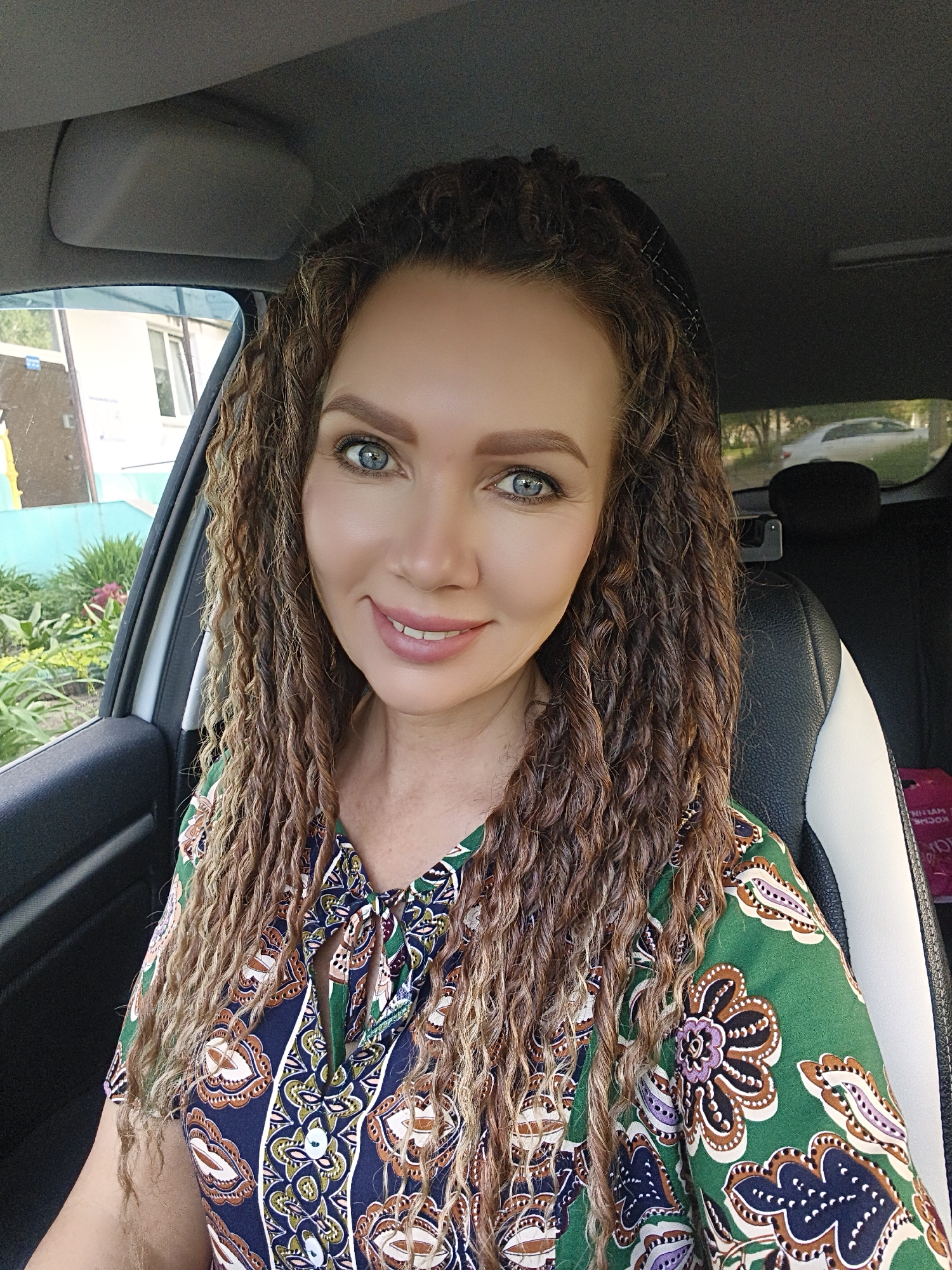 A middle-aged woman is seated in the driver's seat of a car. She is wearing a green and blue patterned dress and has long, curly hair. Her eyes are blue. The car is parked on a street in the city of Unknown, Unknown. In the background is a house with a blue wall and plants. Another car is parked in the distance. The lighting is natural daylight.