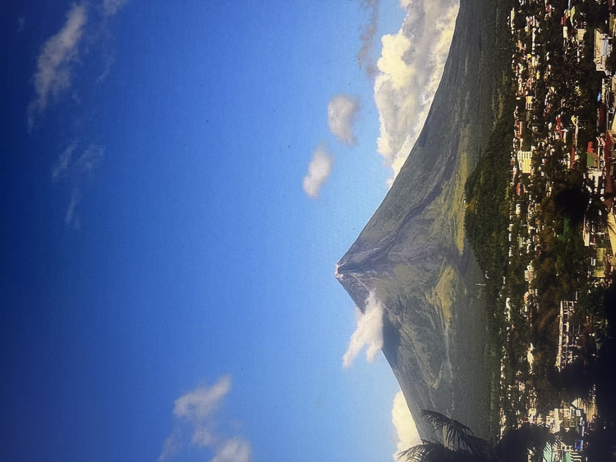 The image shows a mountain with a pointed peak, likely a volcano, adjacent to a city. The sky is blue with scattered clouds. The city is densely populated with buildings of varying colors, mostly red and white. The mountain is green with a visible trail or path leading to the peak. Palm trees are visible in the foreground. The location is not Durmersheim, Germany.