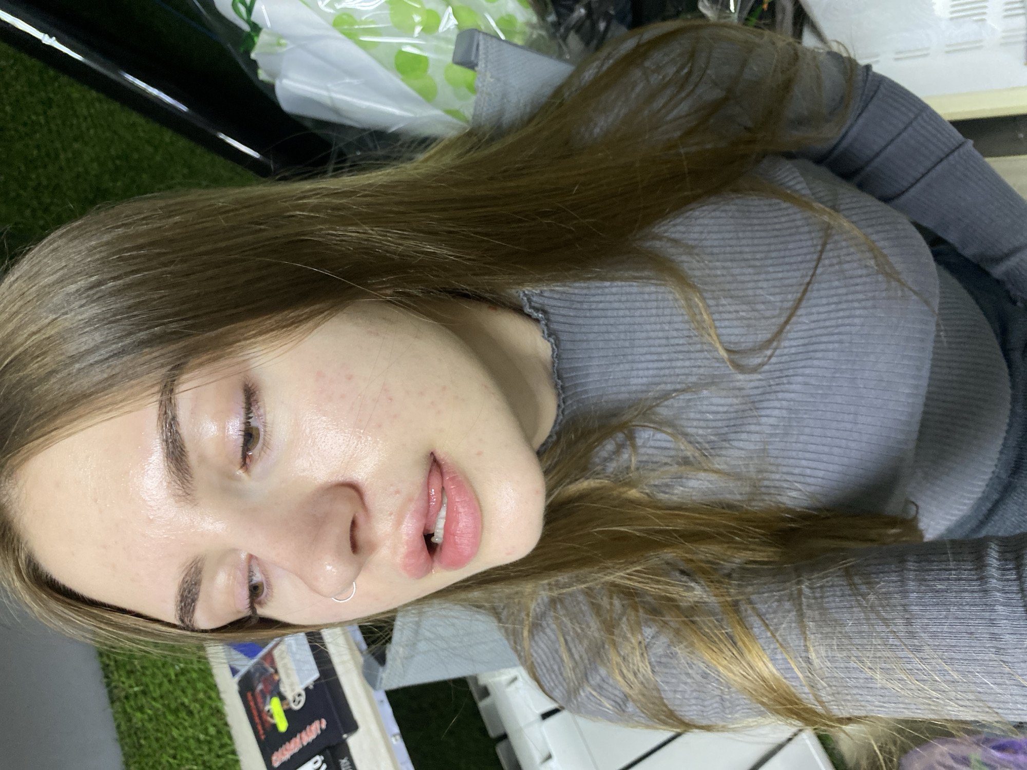 A medium shot is captured from a high angle, focusing on a young woman with long, light brown hair. She is wearing a grey ribbed turtleneck sweater. Her face is turned to the side, with her lips slightly parted and her tongue peeking out. She has a silver hoop piercing in her nose. The background is blurred but appears to be an indoor setting with some greenery and shelves in the distance. The lighting is bright, highlighting the sheen on her skin. The overall impression is a casual, perhaps selfie-style, photograph.