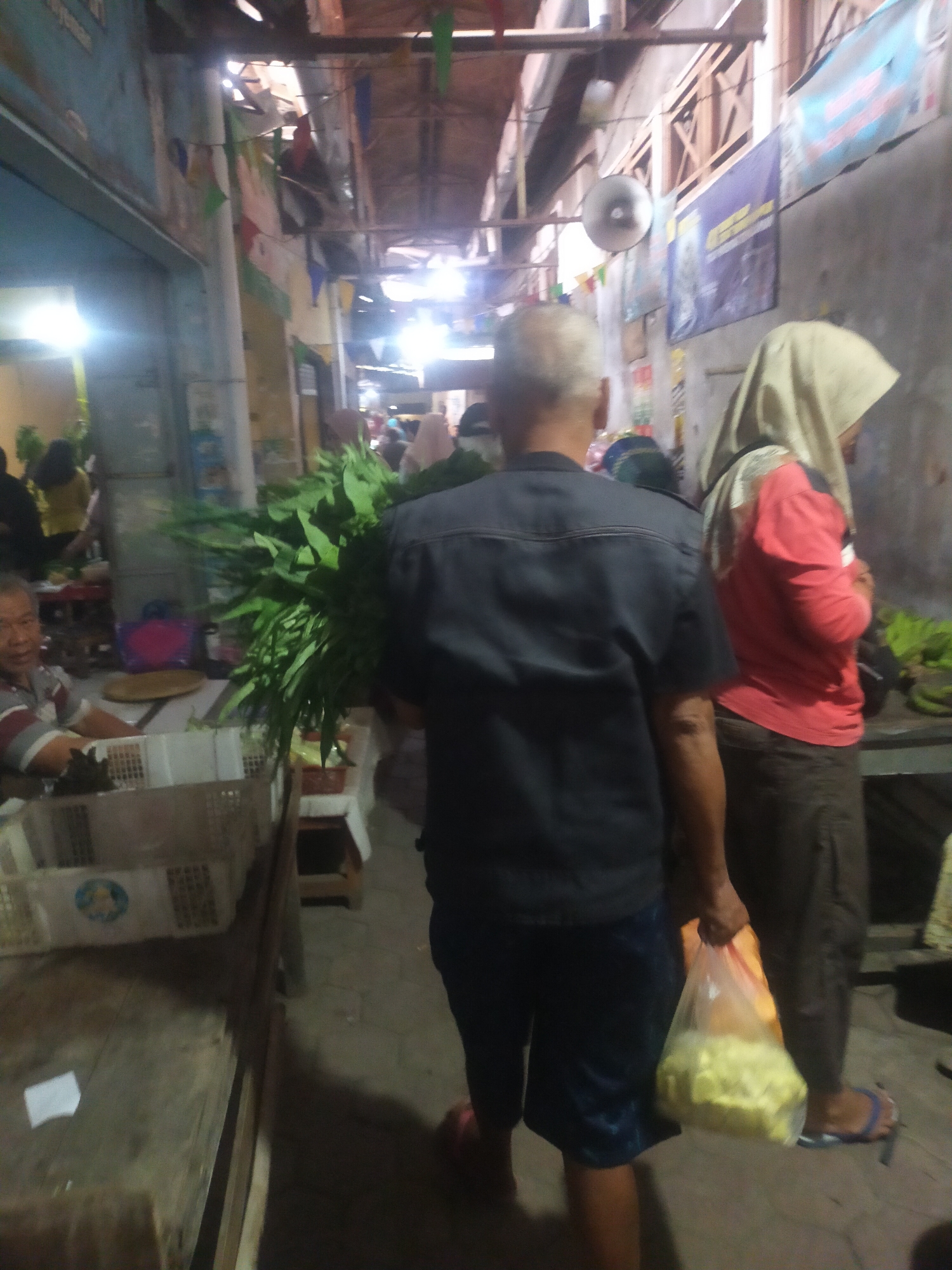 The image depicts an indoor market scene in Trayeman, Indonesia. In the foreground, a man with graying hair is walking away from the camera, carrying a bunch of green leafy vegetables and a plastic bag filled with yellow produce. He is wearing a dark blue short-sleeved shirt and dark blue shorts. To his right, a person wearing a light-colored hijab and a red long-sleeved shirt is walking in the same direction. Further to the left, another man in a striped shirt is visible sitting behind a table with a basket. The market appears to be bustling, with other people and stalls visible in the background. The environment is somewhat dimly lit, with overhead lights and natural light filtering through gaps in the ceiling, suggesting an open-air or semi-enclosed market structure with a wooden roof. Banners and flags are hanging from the ceiling.