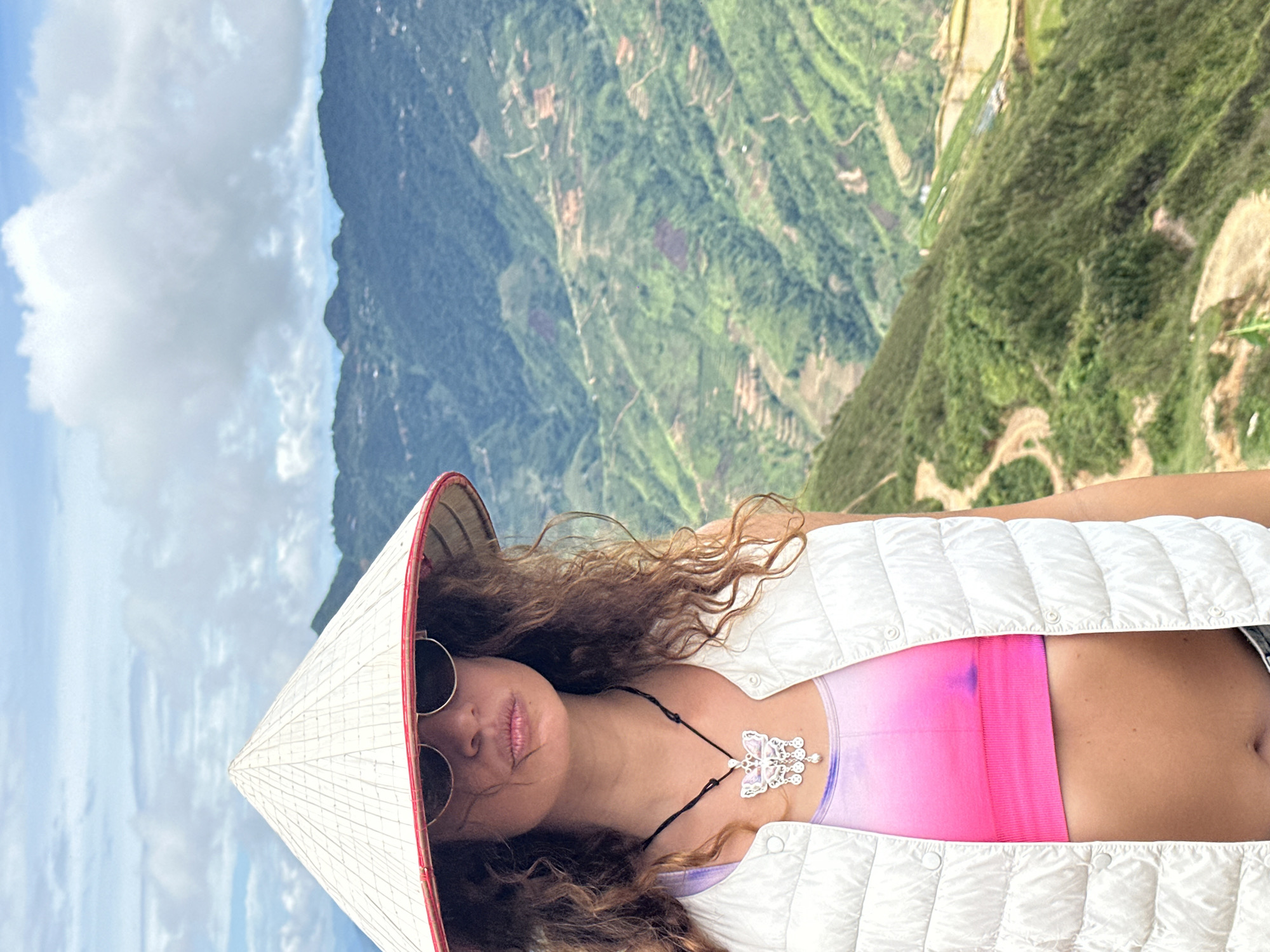 A medium shot captures a woman wearing a conical hat and sunglasses, standing on a mountain slope with terraced fields and lush green vegetation cascading down the hillside under a cloudy sky. She has curly brown hair and is wearing a white puffer vest over a pink and purple tie-dye sports bra, and a silver butterfly necklace. The woman's face is partially obscured by the hat and sunglasses, but her lips are pursed. The background is a panoramic view of the mountainous landscape of Ta Xua, Vietnam, characterized by its distinctive rolling hills and cultivated terraces.