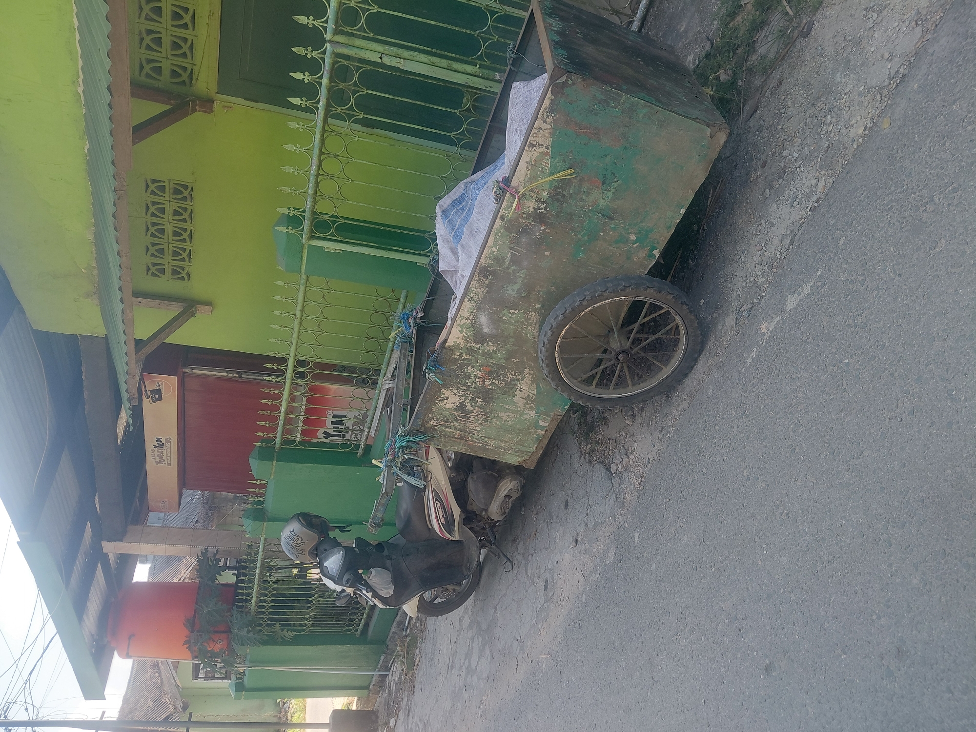 An outdoor, eye-level shot captures a motorcycle with a trailer parked on the side of a paved road in Kendari, Indonesia. The motorcycle, a light-colored automatic scooter, is facing left, with its kickstand down. A person wearing a helmet is seated on the motorcycle, though their face is obscured. The trailer, a weathered green metal box with a single wheel on the right side, is attached to the rear of the motorcycle and contains a large, rolled-up bundle of white and blue fabric.
Behind the parked vehicle is a green metal fence, part of a property line. Beyond the fence, a lime-green building with a corrugated metal awning over a wooden structure is visible. An orange water tank sits on the left side of the building's exterior. The overall scene suggests a residential or commercial area in Kendari. The sky is bright and clear.