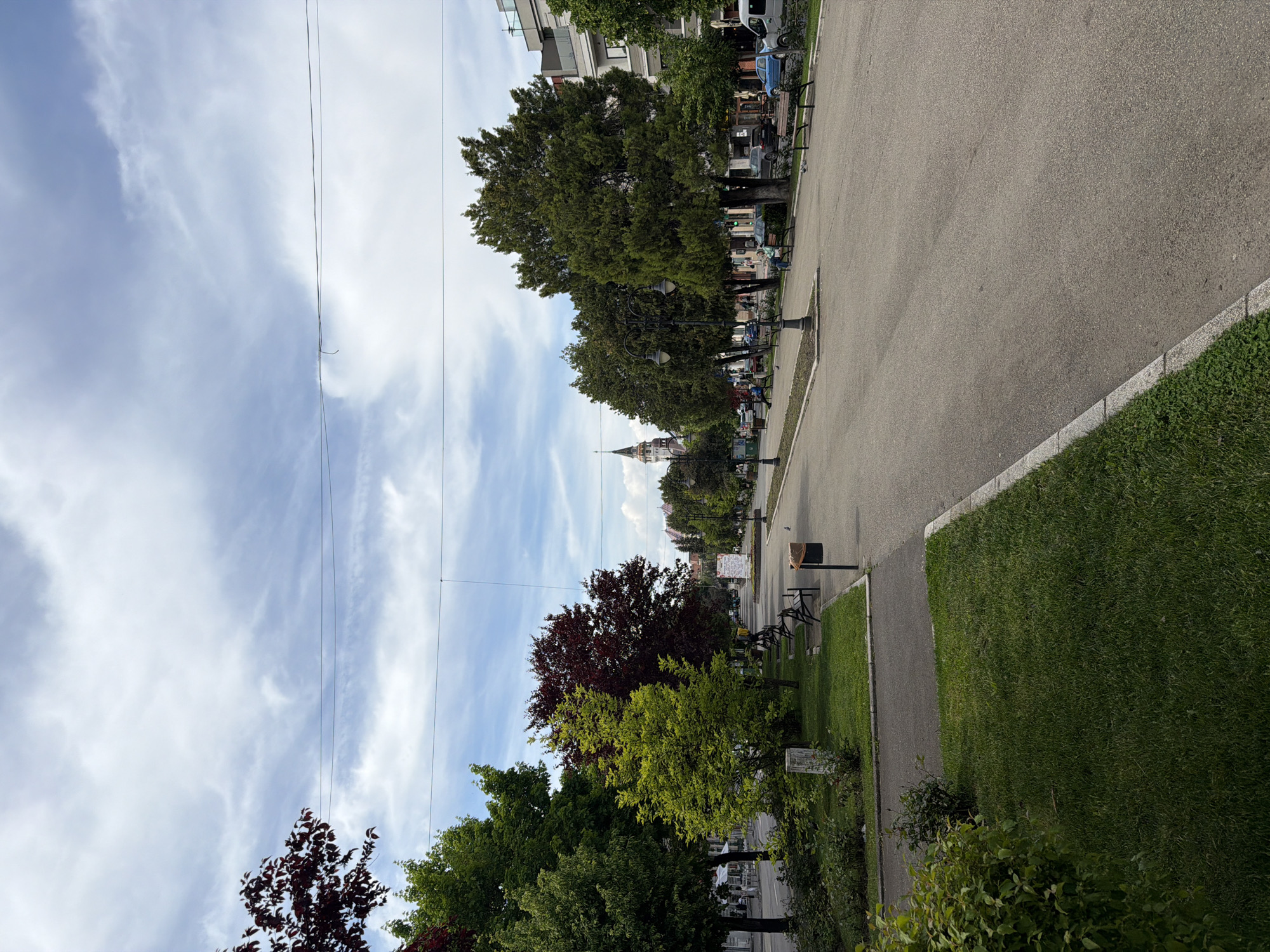 The image shows a park scene in Târgu Mureș, Romania. A wide asphalt path runs lengthwise along the right side of the frame. A strip of grass and a sidewalk border the path on its right, separated by a concrete curb. The left side of the path is lined with trees and benches. In the distance, the silhouette of a tall building or tower is visible. Power lines run diagonally across the sky from the upper left corner. The sky is blue with scattered white clouds.