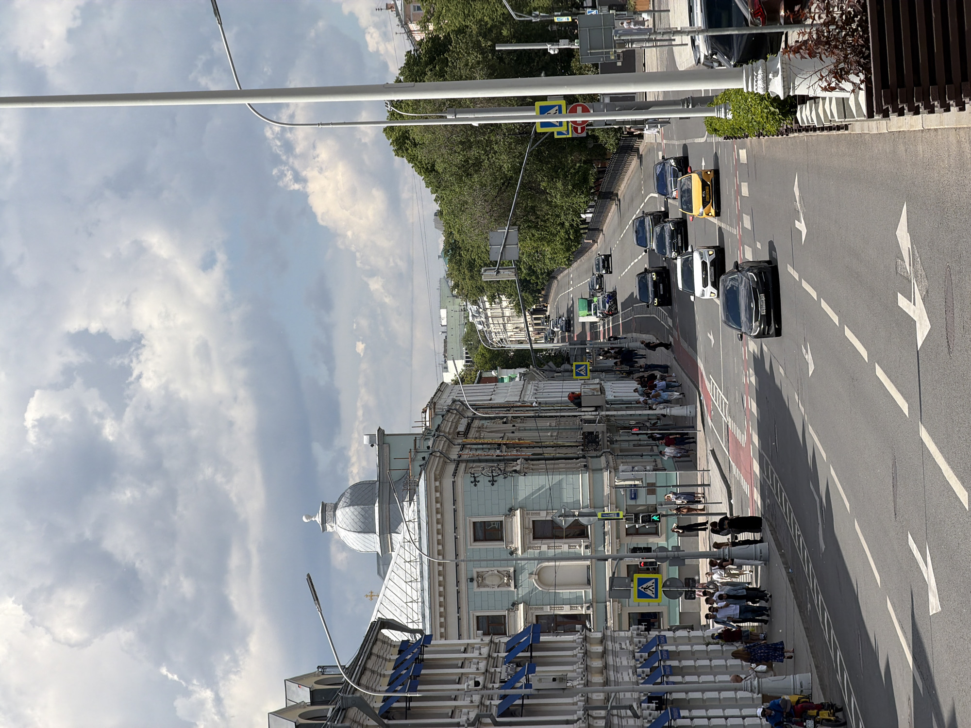 The image depicts a bustling city street in Moscow on a bright, cloudy day. The wide asphalt road, featuring white lane markings and directional arrows, stretches across the middle and right side of the frame. Several vehicles, predominantly dark sedans and black taxis, along with a distinct yellow car, are moving or queued in traffic. A red-painted section, likely a dedicated lane, runs along the right edge of the road.
On the left side of the street, an ornate, light-blue historical building with a prominent silver dome dominates the foreground. Its facade showcases intricate architectural details, multiple windows, and decorative elements. A wide sidewalk in front of this building is densely populated with pedestrians, many gathered at a marked crosswalk. Traffic lights, currently displaying green for vehicles, are visible near the crossing. Further down the street, a line of lush green trees forms a natural boundary, partially obscuring other structures.
The urban infrastructure includes tall, white streetlights and poles supporting traffic signals and overhead cables. Several road signs are clearly visible: a yellow diamond indicating a priority road, a red circular 'no entry' sign, and a blue square 'pedestrian crossing' sign. People on the sidewalk are casually dressed, engaged in daily activities like walking and waiting. No specific emotions are discernible among the crowd. A partial license plate on a black car reads "B399 MP 94". The scene captures the dynamic atmosphere of a typical daytime street in a vibrant city.