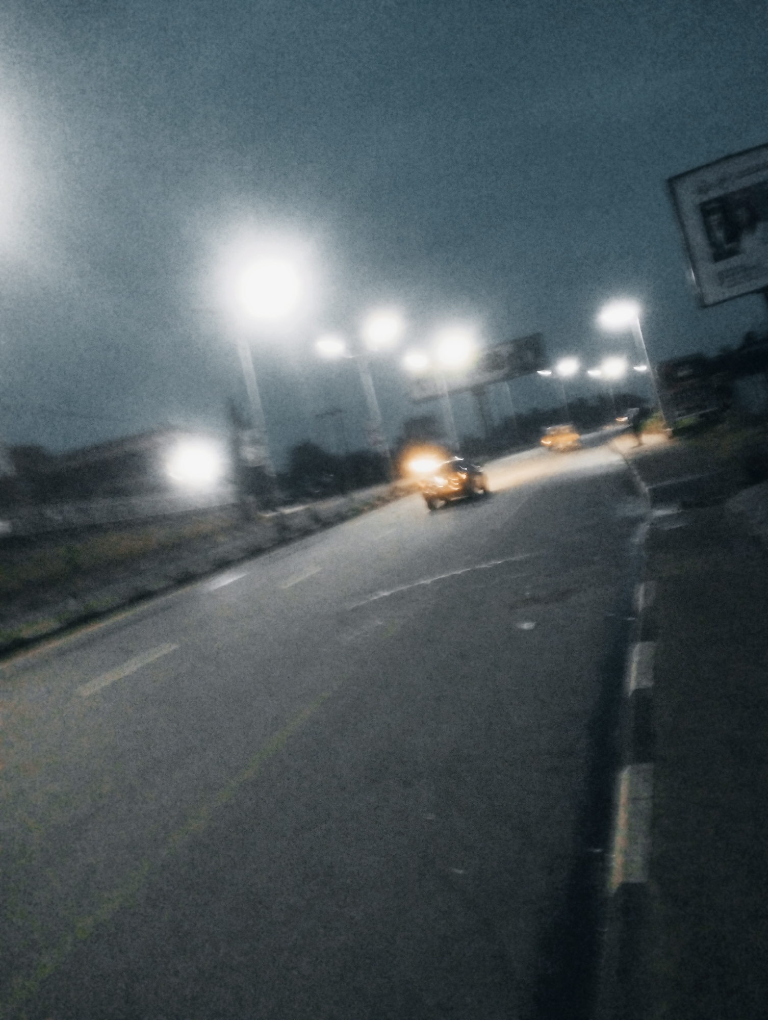 The image depicts a night scene on a street in Jos, Nigeria. The street is paved and appears to be a highway or major road, with streetlights illuminating the scene. There are at least two vehicles visible. One car, appearing to be yellow or orange, is in the foreground, its headlights on, suggesting it is either moving or parked with lights activated. Another vehicle, also yellow or orange, is further down the road. A figure, possibly a person, is standing near the second vehicle. The environment includes a roadside verge with some vegetation. A billboard or signpost is visible in the background. The overall atmosphere is dark, with the streetlights creating bright spots and casting long shadows, contributing to a somewhat blurred and impressionistic view due to low light conditions and potential motion blur.