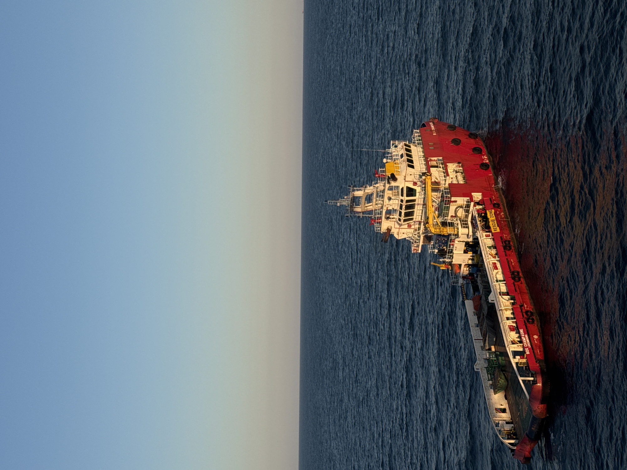 An offshore supply vessel, named "BOS CHAMPAGNE LUXEMBOURG", is seen at sea under a clear sky. The vessel is predominantly red with white and yellow superstructure. It has a large open deck area at the rear, carrying some equipment. The water is a deep blue, with gentle waves reflecting the sunlight. The horizon is visible in the distance, with a faint haze. The image captures the vessel from a slightly elevated angle, showcasing its size and its presence in the open sea. There are no people visible on the deck or in the superstructure. The overall environment suggests an industrial maritime setting.