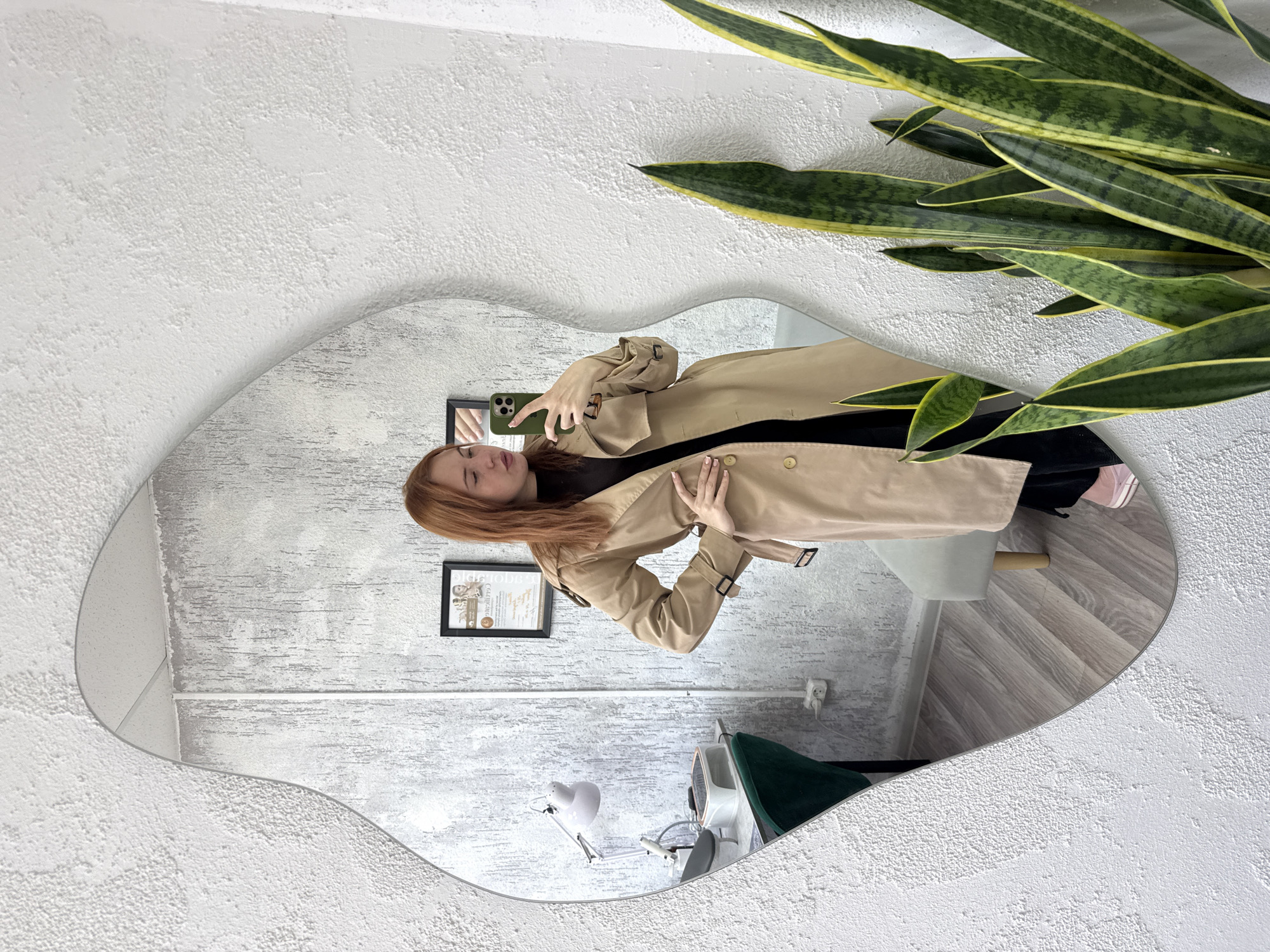 A person is seen in a mirror selfie, captured in an irregularly shaped (wavy-edged) mirror, which is mounted on a textured white wall. The subject, a woman with reddish-brown hair, is wearing a beige trench coat over a dark top. She holds a dark green smartphone (likely an iPhone due to the camera configuration) in her right hand, aiming it towards the mirror, while her left hand rests casually on her waist.
The background, visible both directly and reflected in the mirror, consists of a white textured wall. Parts of the wall visible in the mirror have a different, possibly smoother, greyish texture. On the right side of the mirror, a tall, green and yellow variegated snake plant (Sansevieria) is partially visible. The reflection in the mirror also shows a framed document or certificate hanging on the wall behind the person; some faint text like "ДОБ" or similar is barely discernible. A white electrical outlet is also reflected on the wall. Further down, the mirror reflects parts of the room's interior: a dark green upholstered chair, a white stool or table with wooden legs, a white articulated task lamp, and a light-colored wooden floor with a chevron or herringbone pattern. The overall setting appears to be an indoor space with modern decor, possibly a salon, office, or stylish home, during daylight hours.