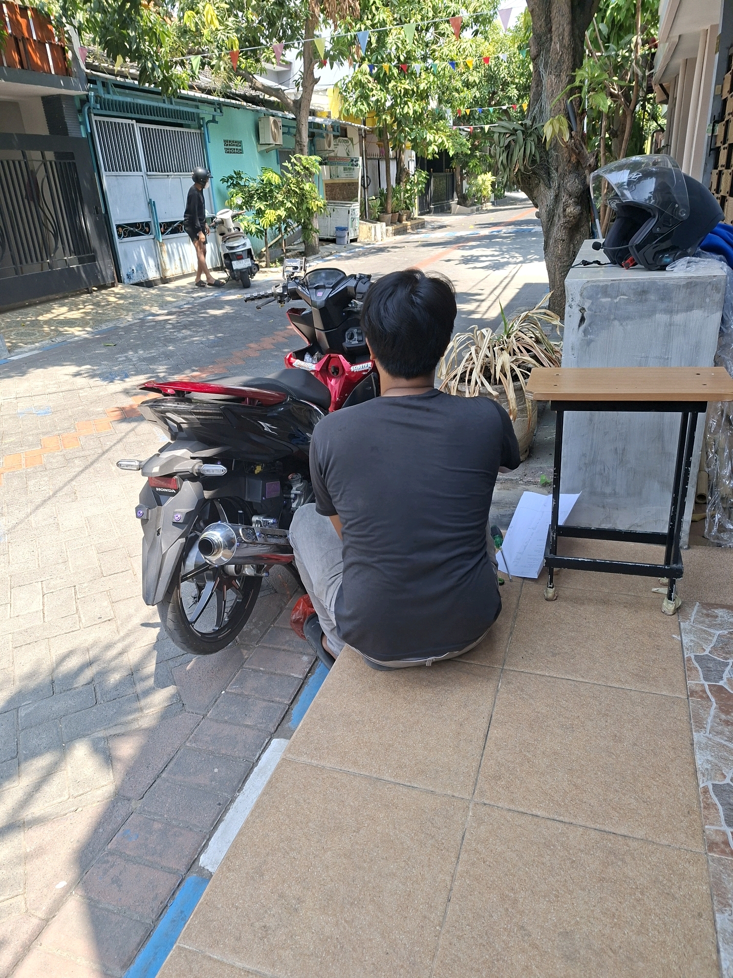 In Surabaya, Indonesia, a person is sitting on a tiled patio next to a red and black motorcycle. The person, viewed from behind, is wearing a dark t-shirt and light-colored pants. The motorcycle is parked on a paved street. In the background, there is a building with a turquoise facade, a metal gate, and trees with colorful flags strung between them. A black helmet rests on a concrete block near a small wooden table with a metal frame. The scene appears to be on a sunny day, casting shadows on the ground.