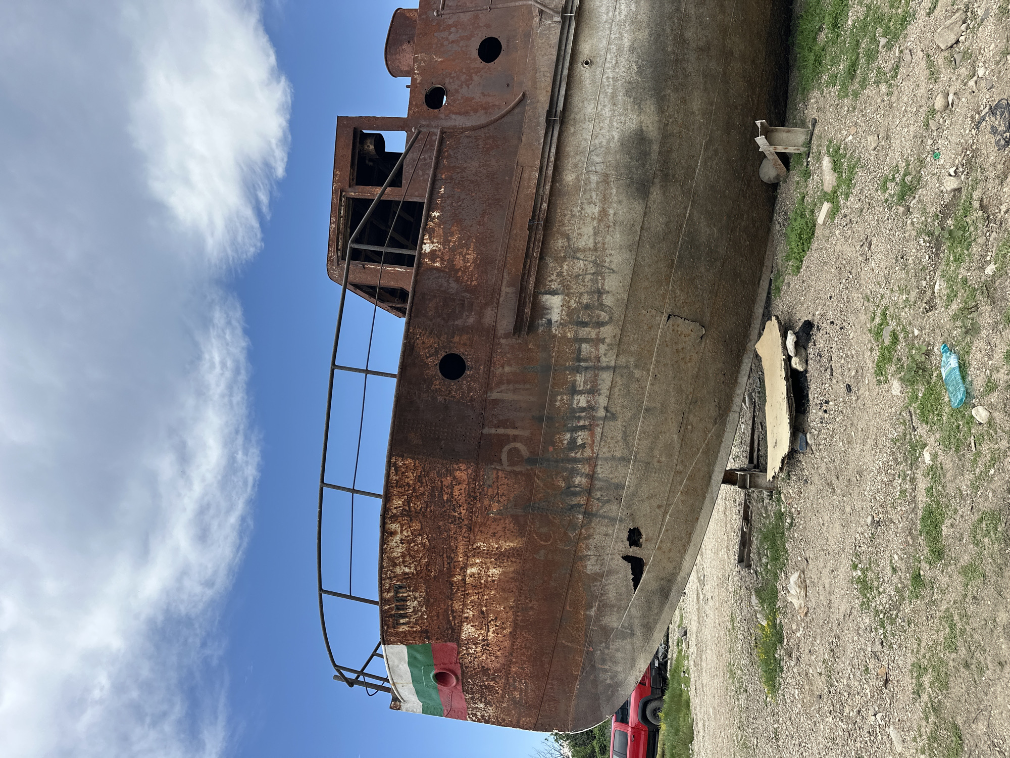 The image captures a severely rusted and derelict metal boat, dry-docked on an unpaved lot in Shiroko pole, Bulgaria, under a partly cloudy blue sky. The vessel exhibits advanced corrosion, with the upper hull and superstructure covered in a reddish-brown rust, transitioning to a lighter, oxidized grey on the lower hull. The bow displays remnants of a painted Bulgarian flag, identified by horizontal white, green, and red stripes. Multiple circular openings, consistent with portholes, are visible on both the hull and the exposed framework of the superstructure, which is devoid of windows or doors. Black graffiti in Cyrillic script is present on the rusted hull. The boat is supported by various makeshift blocks and stands. The surrounding ground consists of gravel, dirt, and sparse patches of grass, with discarded plastic bottles and other debris scattered across the surface. A red pickup truck is partially visible parked in the background to the left. No people are present in the scene.