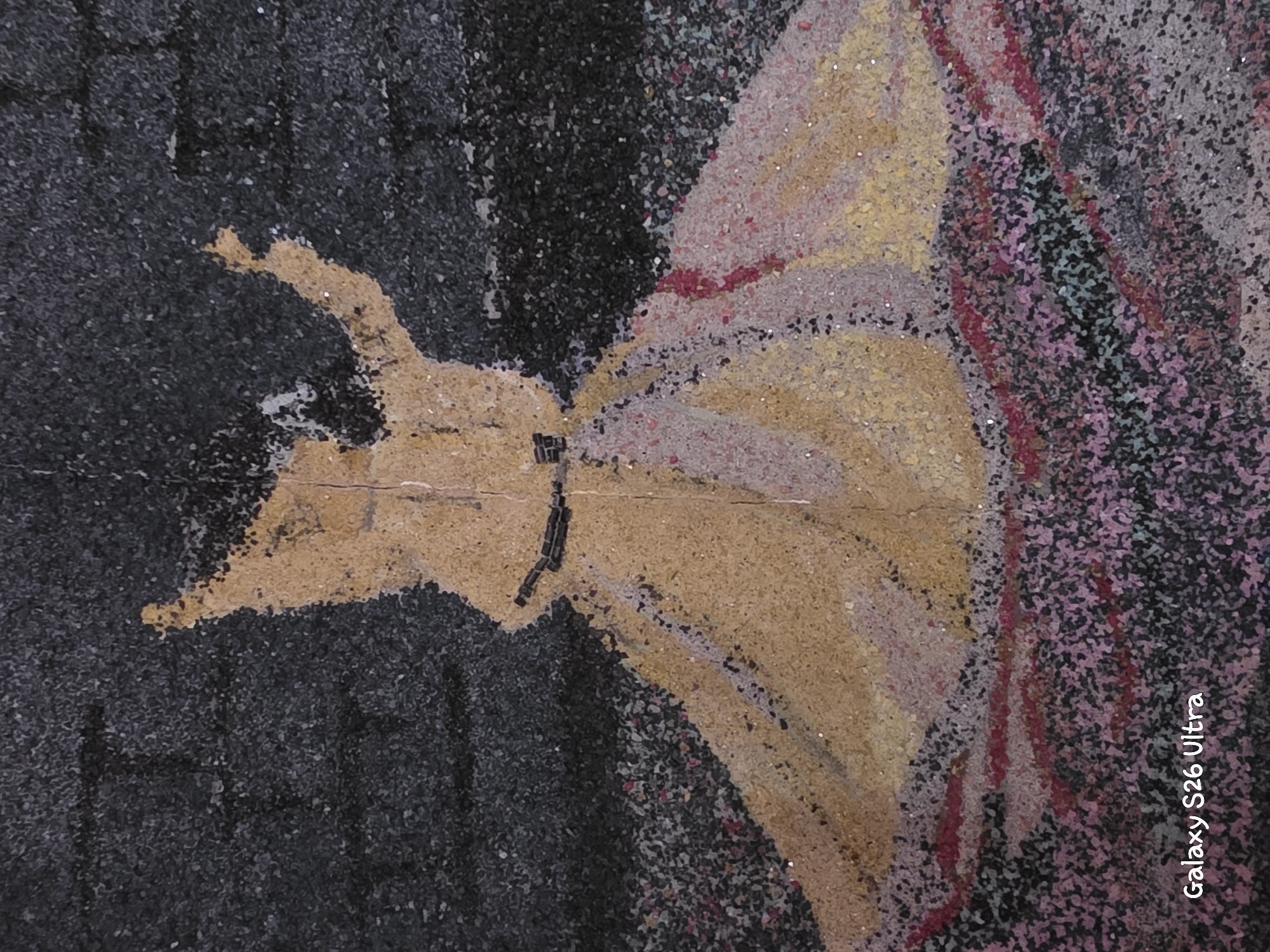 The image shows a mural painted on a rough, textured surface, possibly a brick wall, in Plovdiv, Bulgaria. The mural depicts a figure in a flowing yellow robe, possibly a dervish, in mid-twirl. The background is dark gray with a textured, brick-like appearance. The figure's robe is spread out, with shades of yellow, pink, and red creating a sense of movement and vibrancy. The lower portion of the mural features a pattern of various colors, including red and purple. The mural is somewhat abstract, with the details of the figure rendered in a coarse, granular style. The lighting suggests it is daytime, with no direct sunlight casting harsh shadows. The overall impression is of an artistic representation of a dynamic dance or performance.